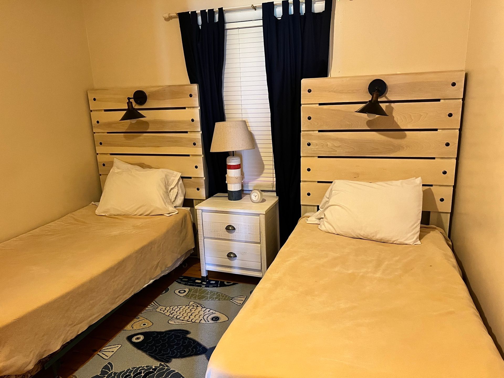 Small bedroom with two single beds, a nightstand, lamp, and window with dark curtains.