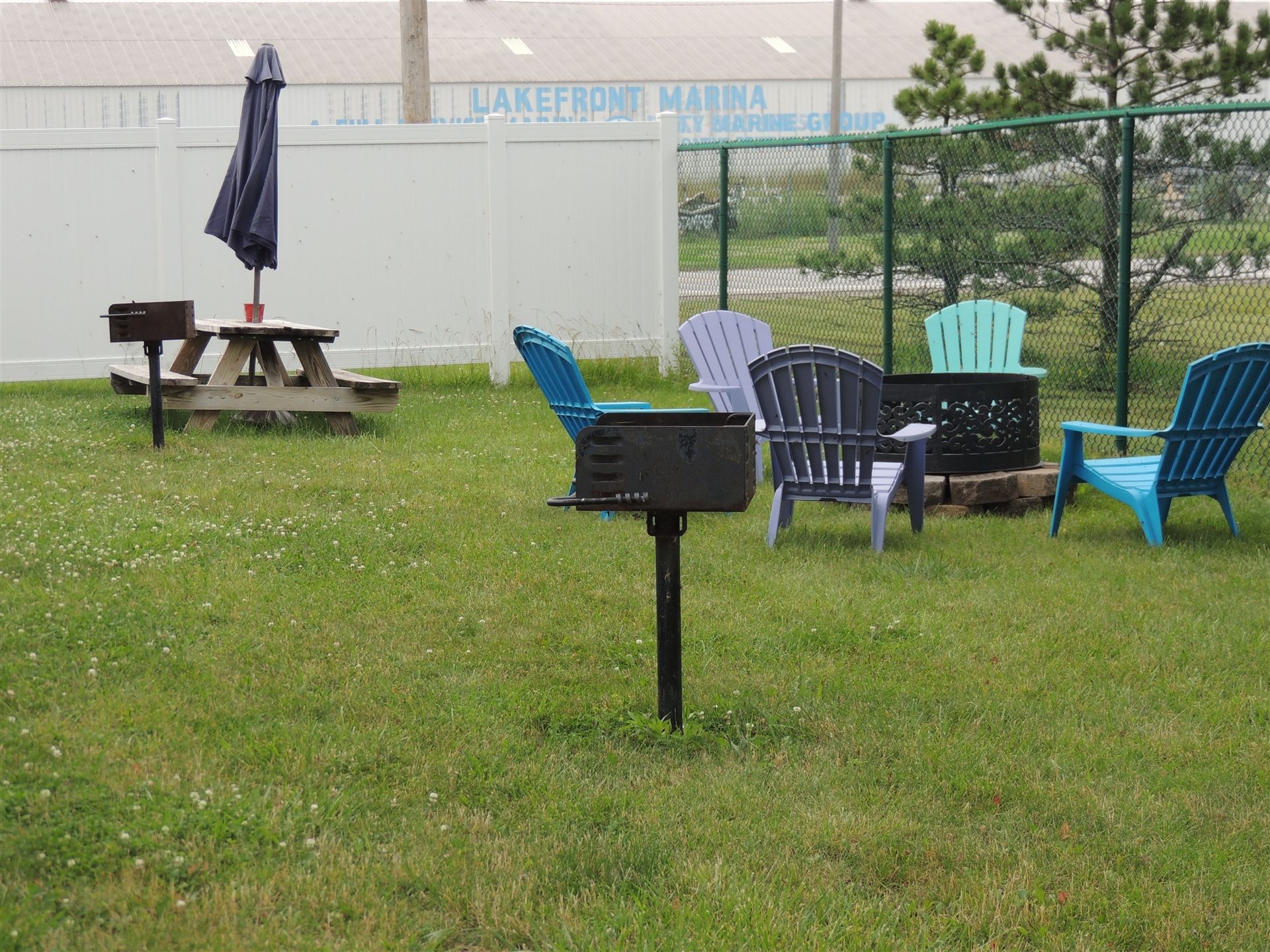 Grassy park area with picnic table, grill, and colorful chairs around a fire pit under a cloudy sky