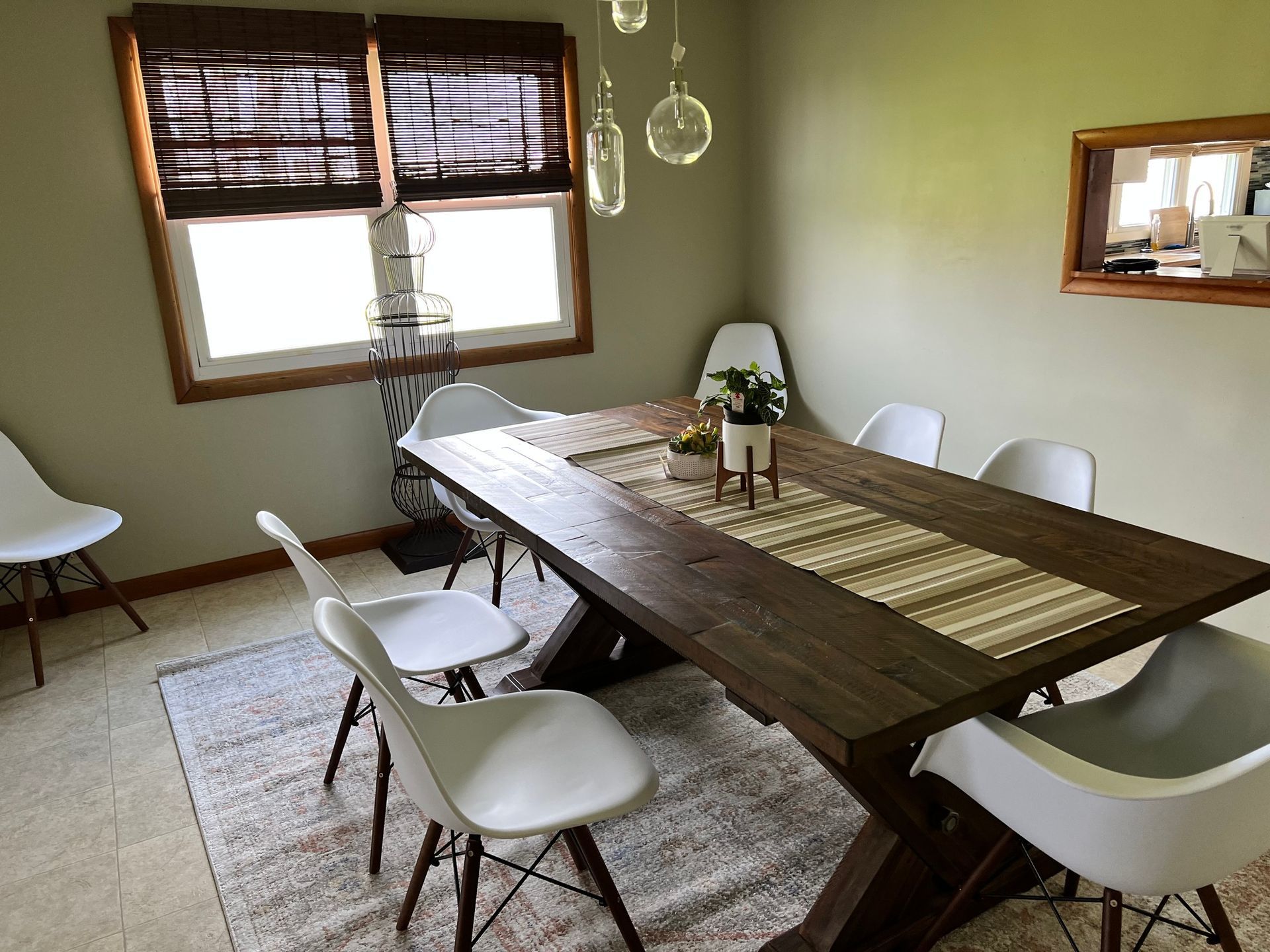 Dining room with a long wooden table, white chairs, and a window with brown blinds.