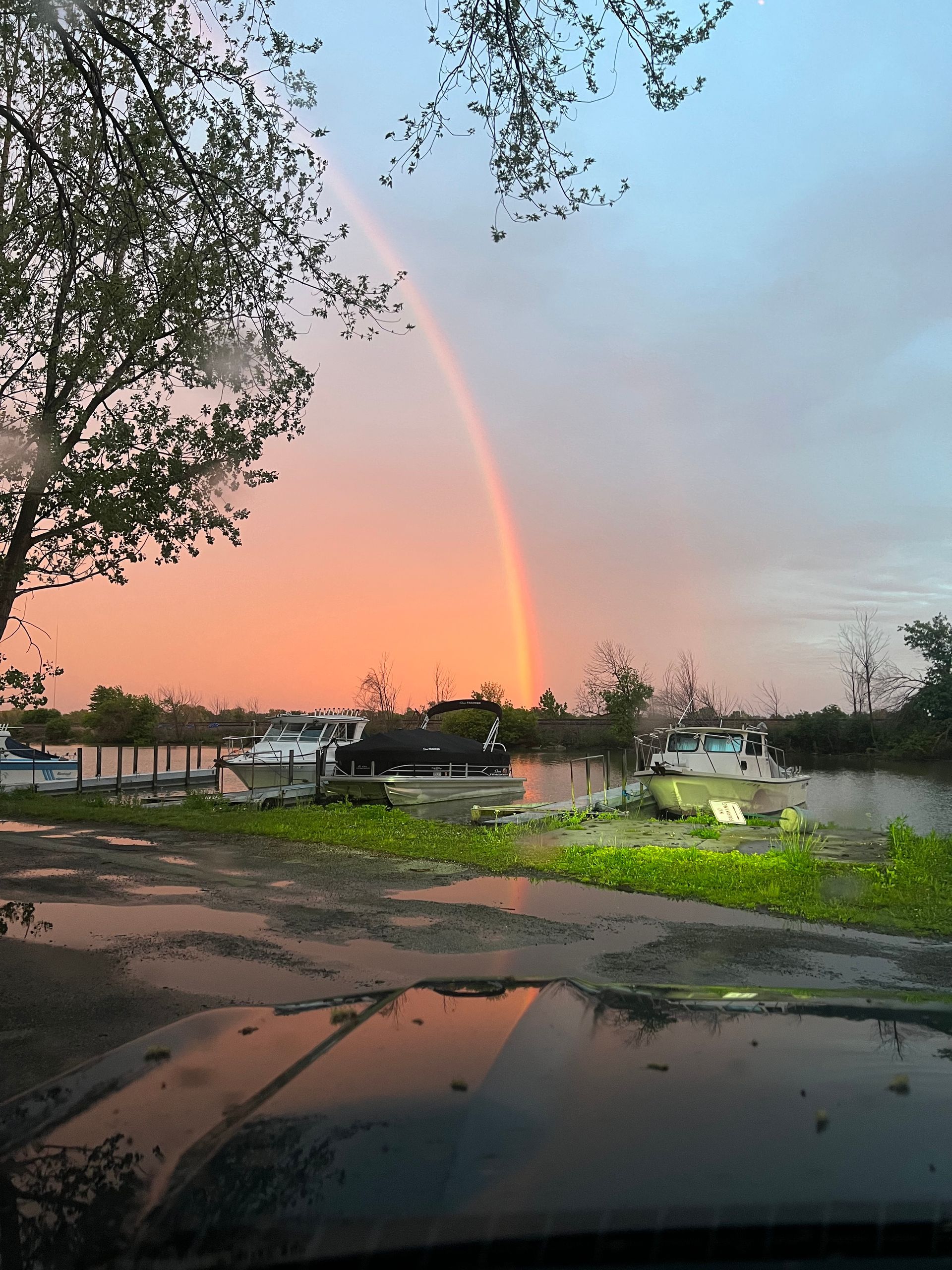 Rainbow over a parking lot at dusk, seen through a car windshield with trees and parked cars.