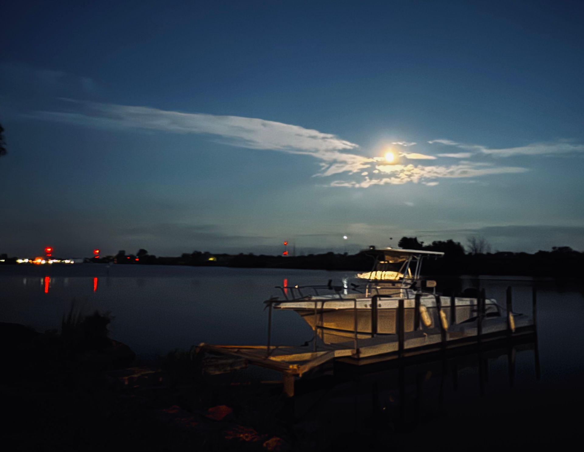 Moonlit lake with a dock and boat silhouettes, city lights reflecting on the calm water at dusk