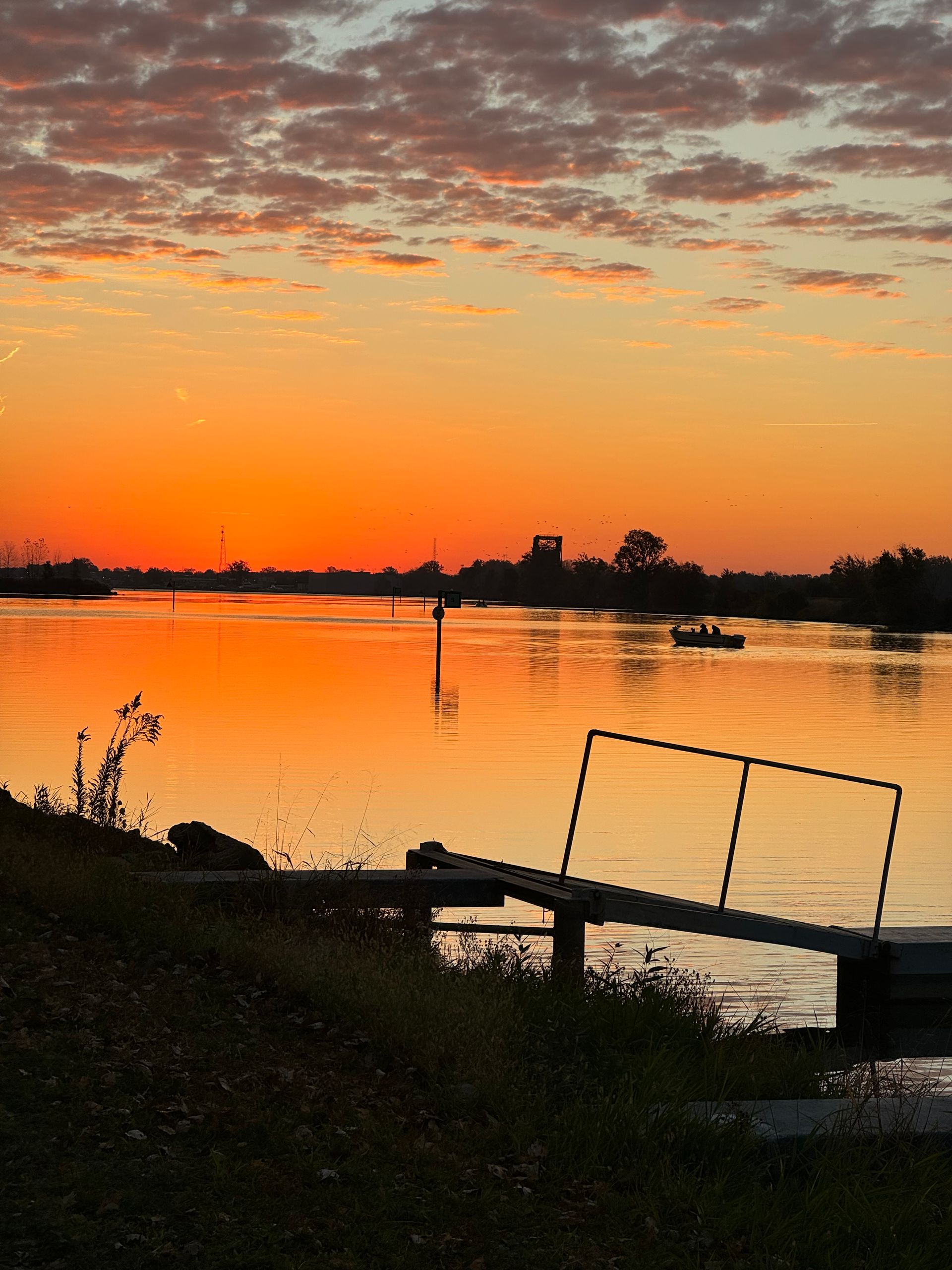Sunset over a calm river with orange sky, silhouetted trees, and a small dock in the foreground