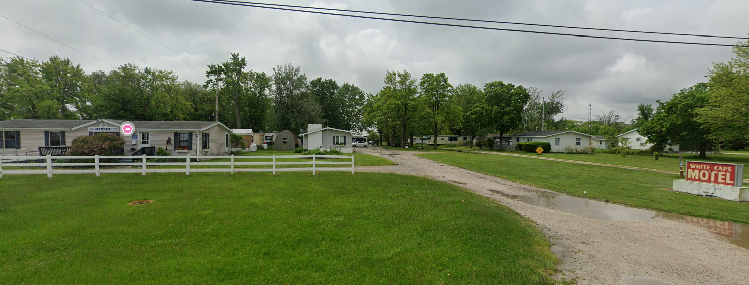 Rural roadside with white fence, grassy field, gravel driveway, and small building under cloudy sky