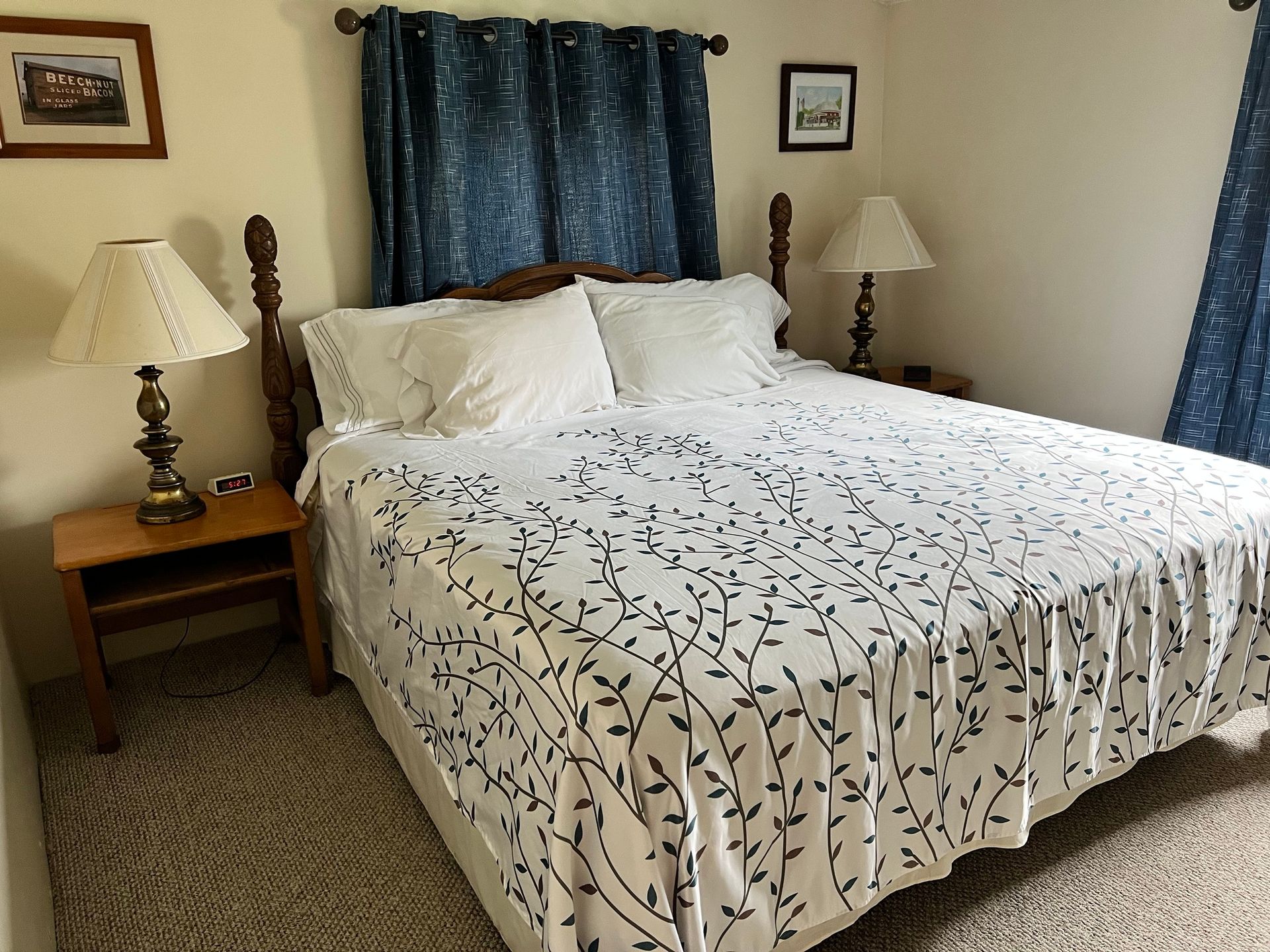 Bedroom with a neatly made bed, blue curtains, two bedside lamps, and framed wall art.