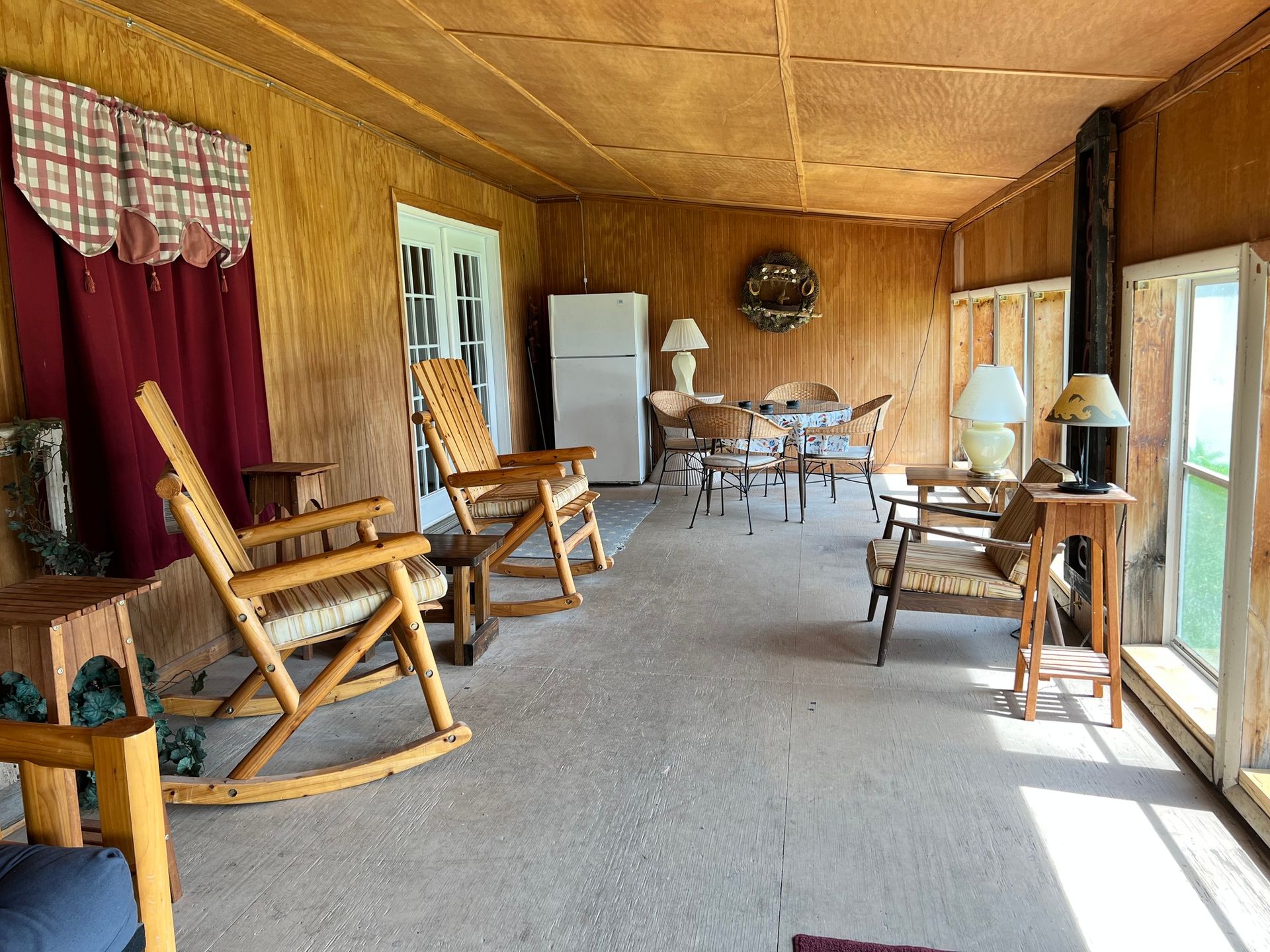 Wood-paneled porch with rocking chairs, small table, lamps, and a refrigerator along the wall