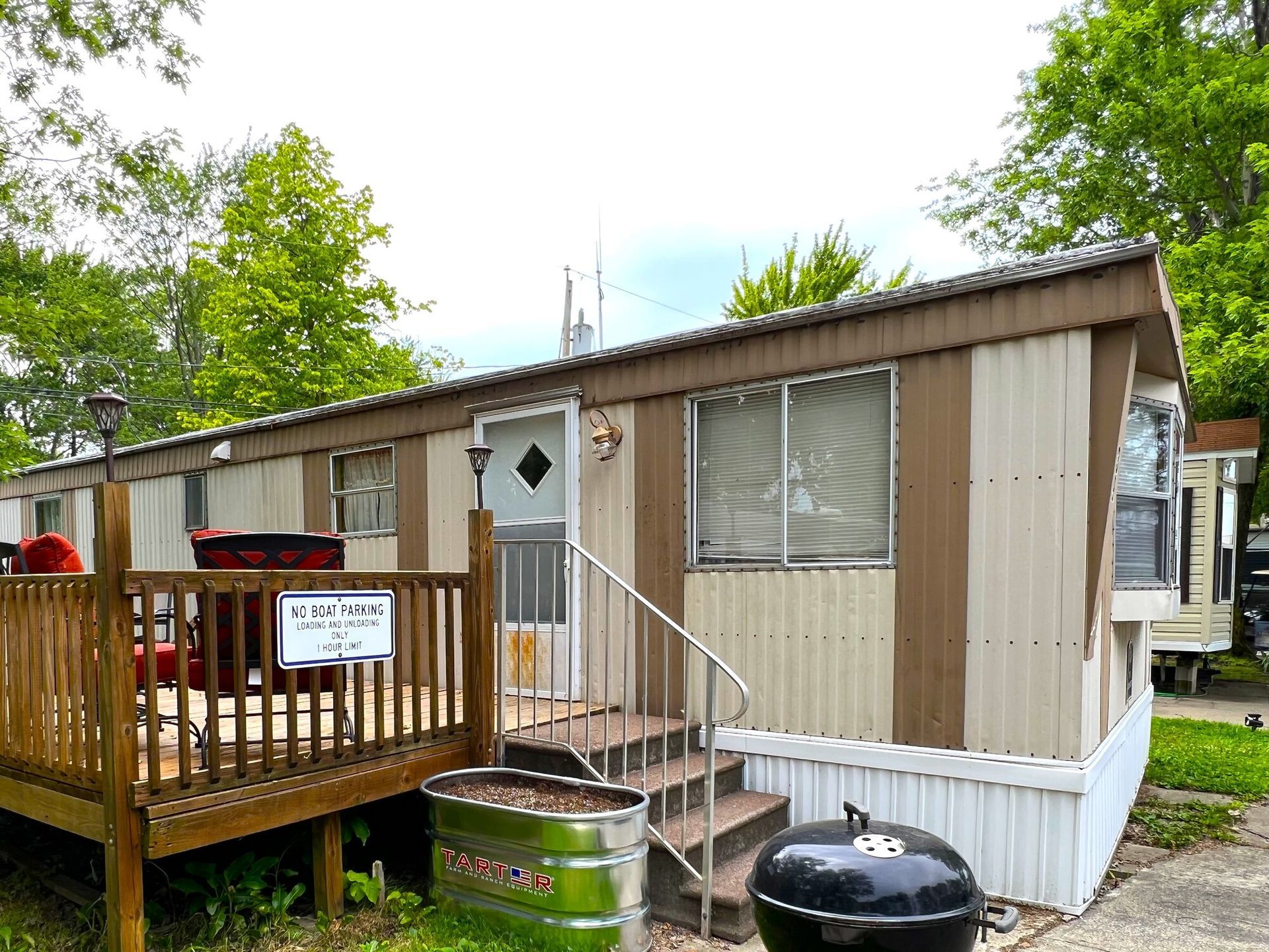 Small beige mobile home with a wooden porch, steps, and black grill in a leafy yard.