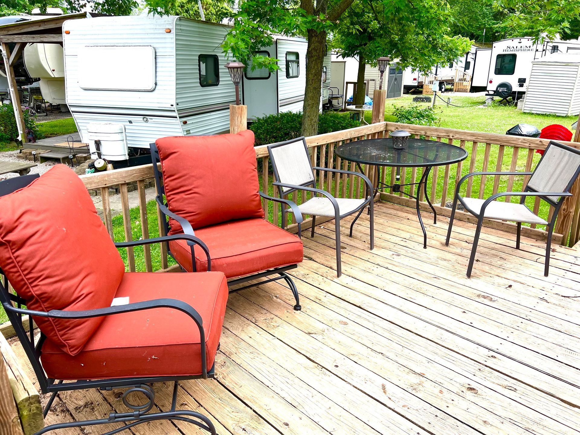 Patio with red cushioned chairs and a round table on a wooden deck beside parked RVs and trees