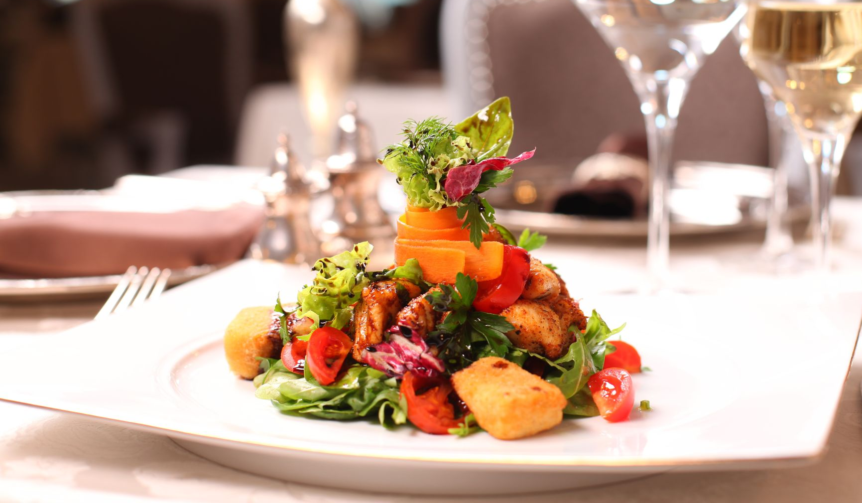 Gourmet vegetable salad on a white plate at a formal dining table, with blurred wine glasses in the background