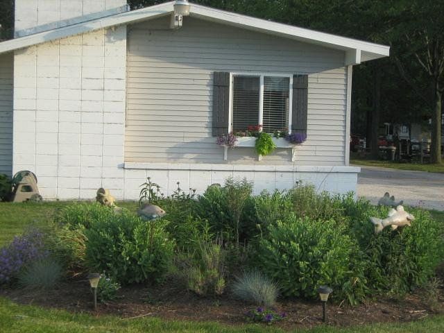 Small white house with gray shutters and flower beds in front, beside a road and trees