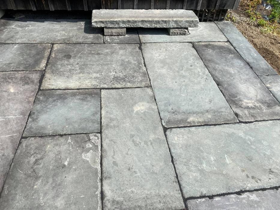 Gray stone patio with a small stone bench in front of a wooden structure.