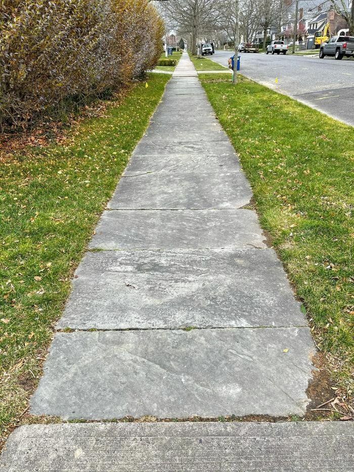 Sidewalk through residential area, bordered by grass and bushes. Pavement shows signs of wear.