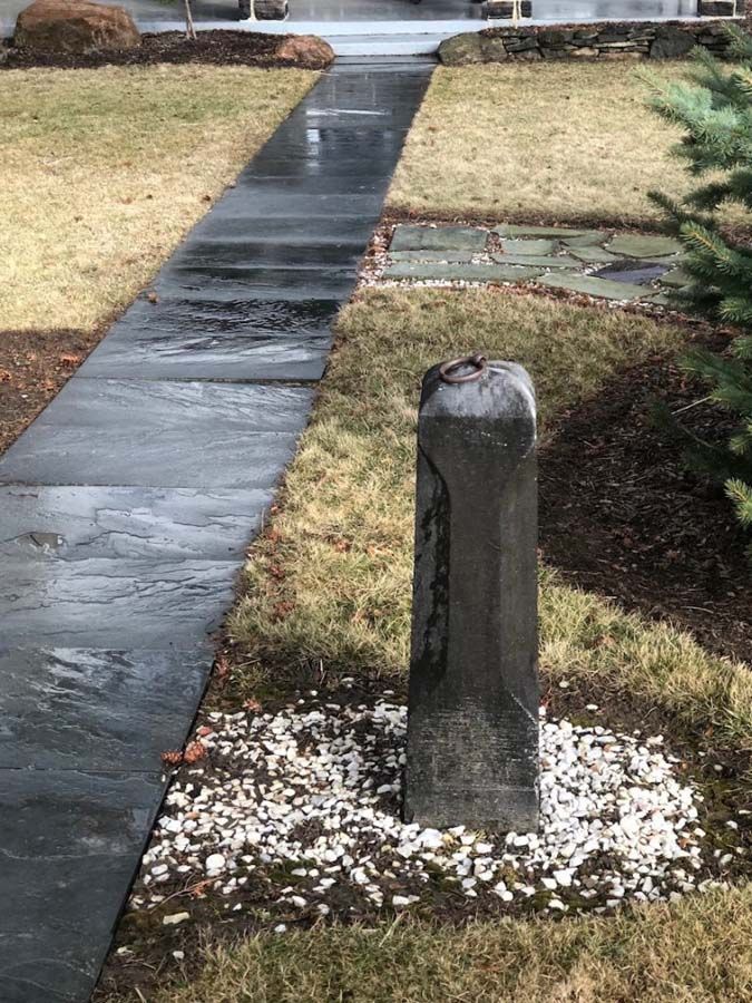 Stone post in a bed of white gravel, next to a wet paved walkway and lawn.
