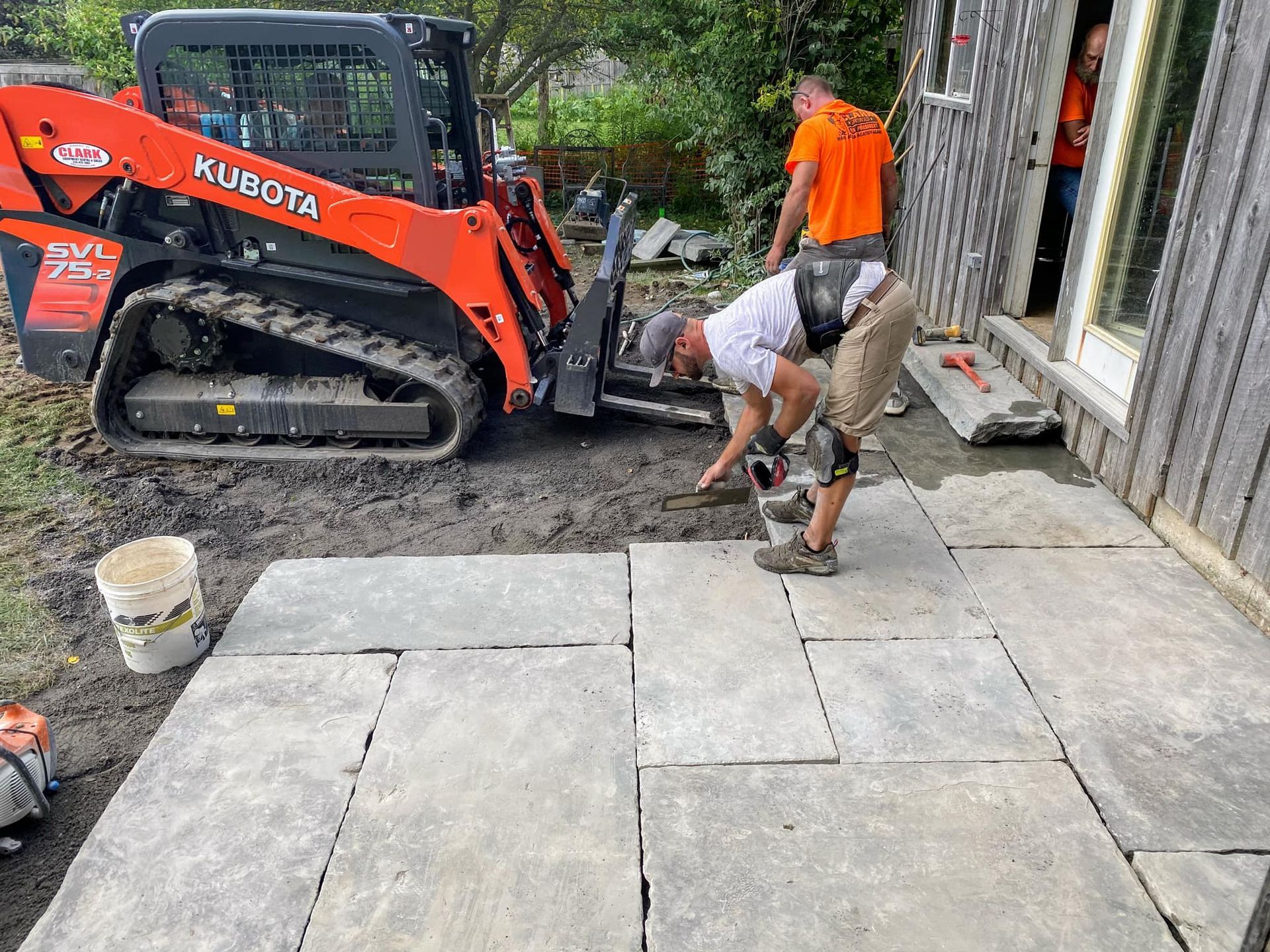 Workers laying stone patio with Kubota skid steer; outdoor setting.