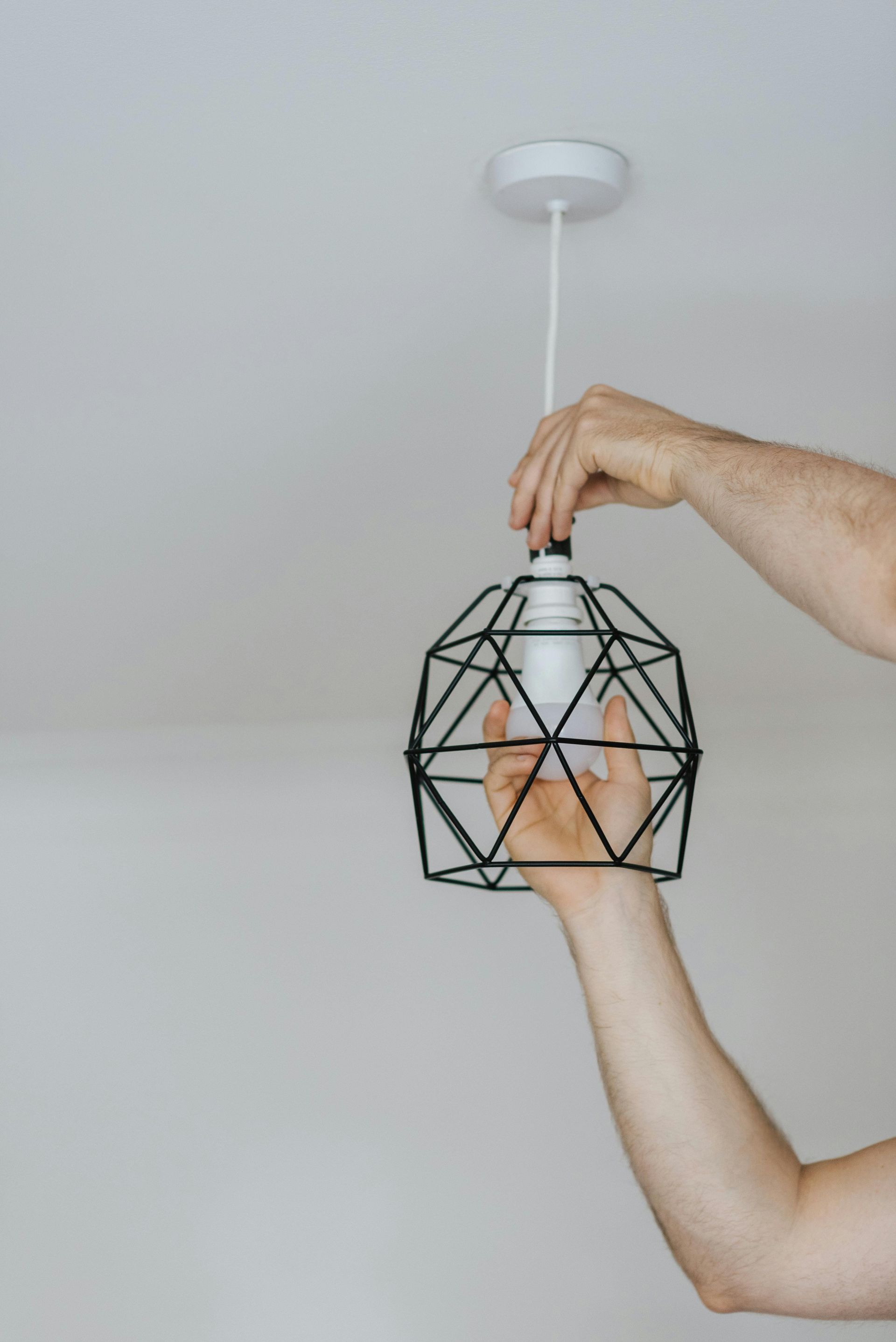 Person installing a lightbulb in a black geometric pendant lamp, against a white ceiling.