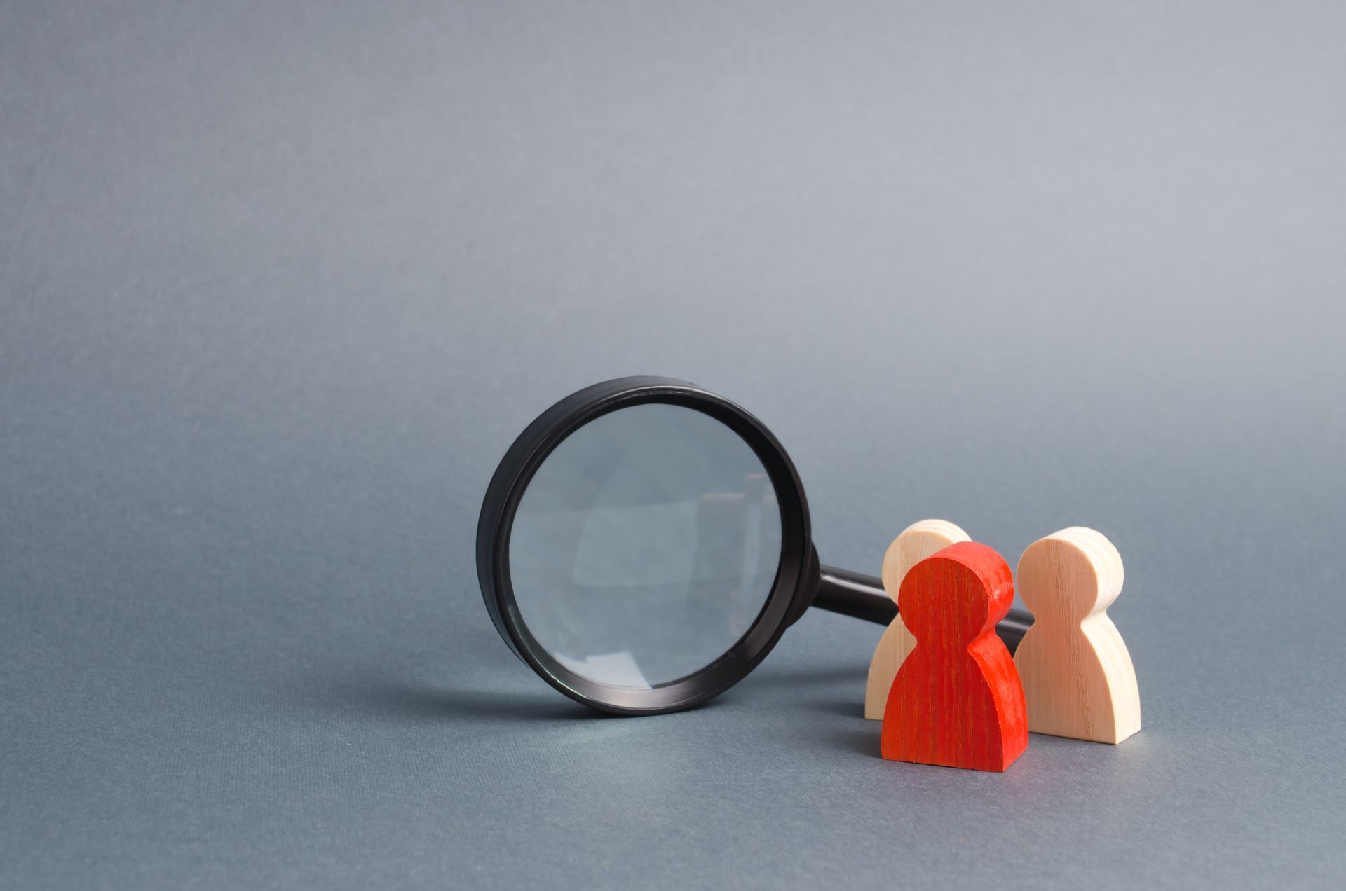 Magnifying glass over three wooden figures, one red, on a gray background.