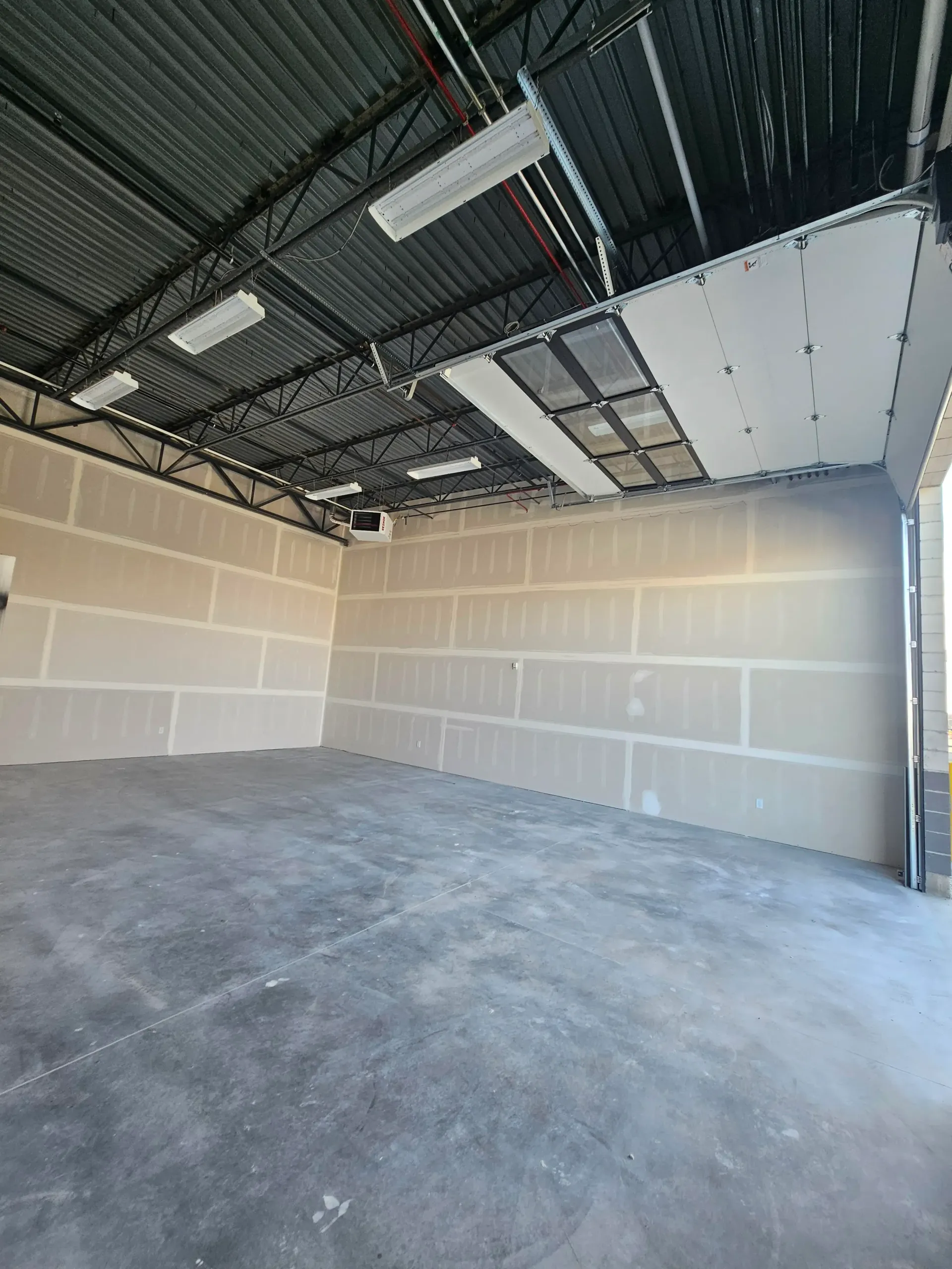 Interior of an unfinished commercial space: concrete floor, drywall walls, metal ceiling structure, open garage door.