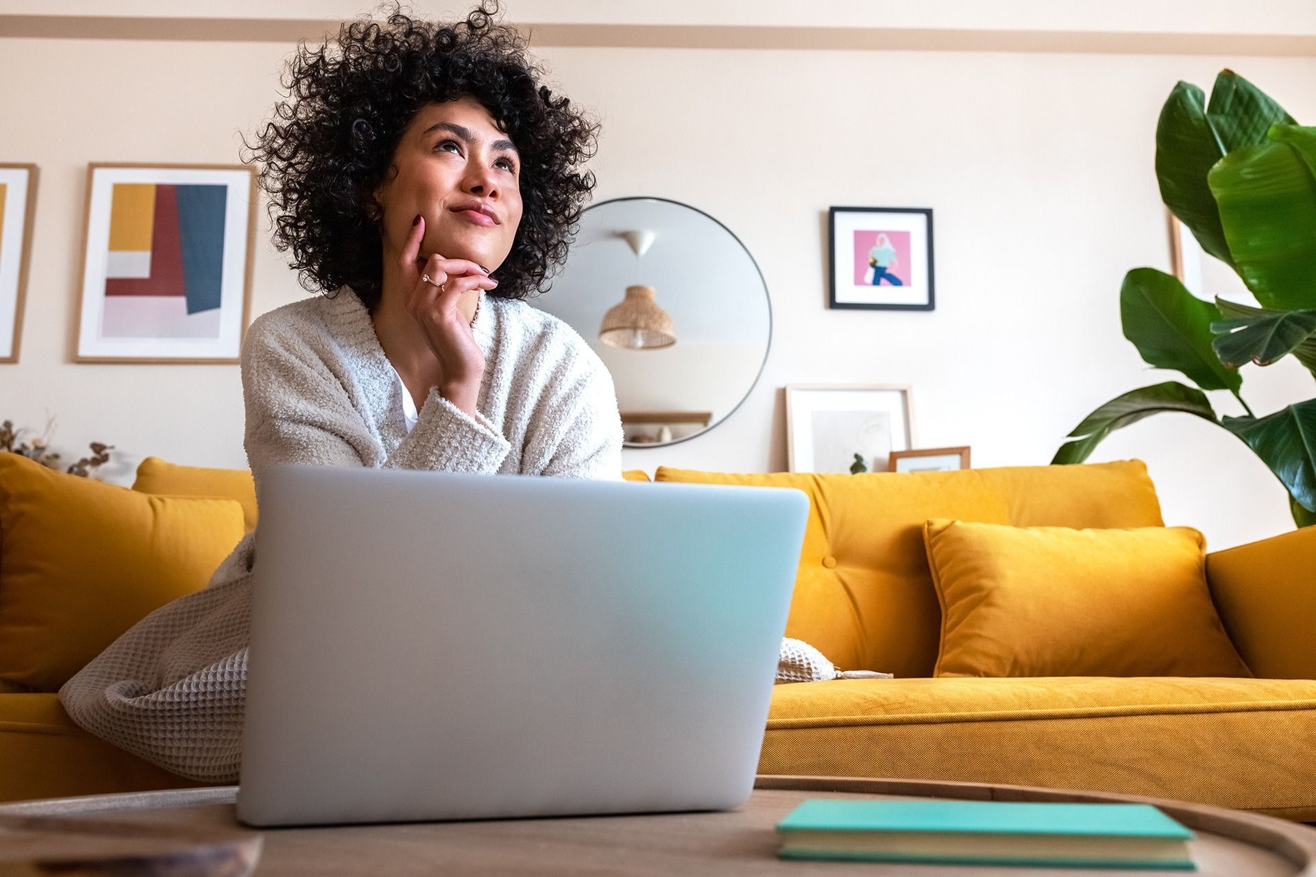 Woman on yellow couch, laptop open, looking up thoughtfully in living room with art and plant.