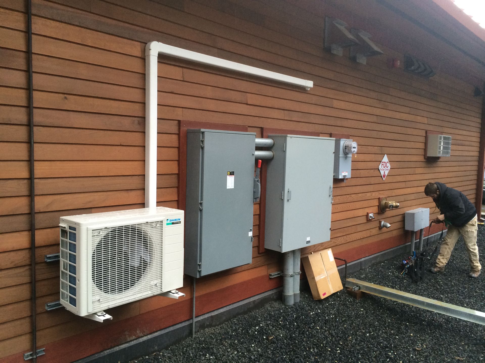 Outdoor utility equipment and air conditioners mounted on a wooden wall, with a worker crouching nearby.