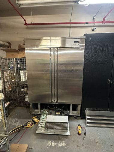 Stainless steel commercial refrigerator in a utility room, with tools and supplies on the floor.