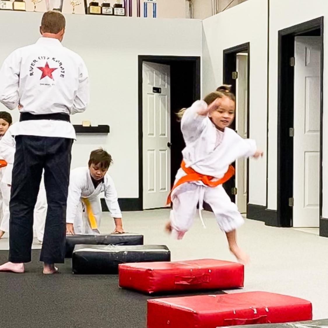 a man in a white karate uniform with a star on the back is watching a little girl jump over red blocks