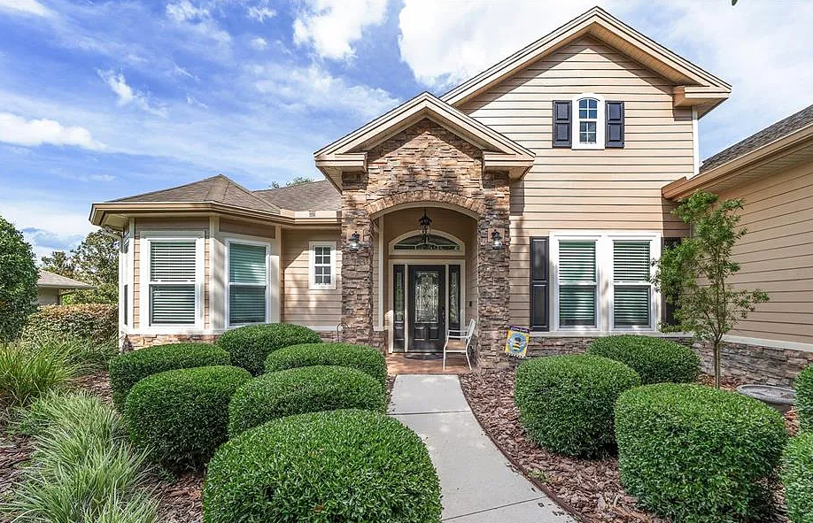 Beige house with stone facade entry, arched doorway, black shutters, and well-manicured bushes along the path.