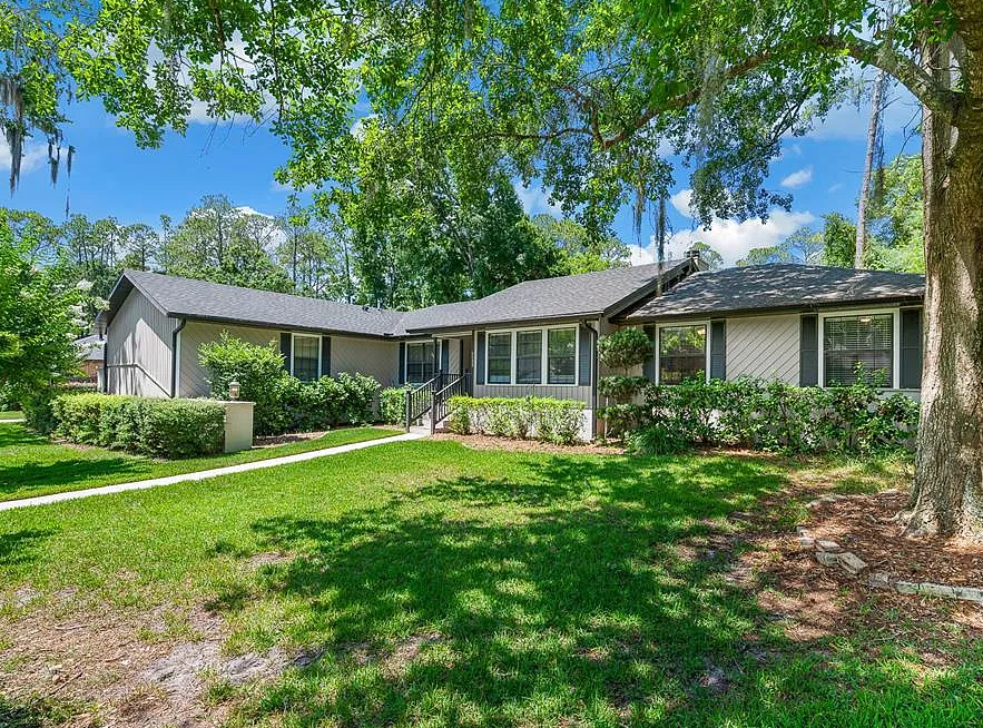Beige house with stone facade entry, arched doorway, black shutters, and well-manicured bushes along the path.