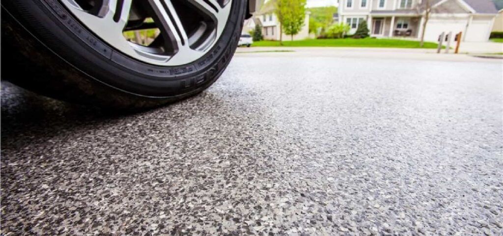 Car tire on a freshly paved gray driveway in front of a suburban house.