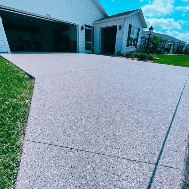 Gray speckled concrete driveway in front of a white house with an open garage door and green grass.