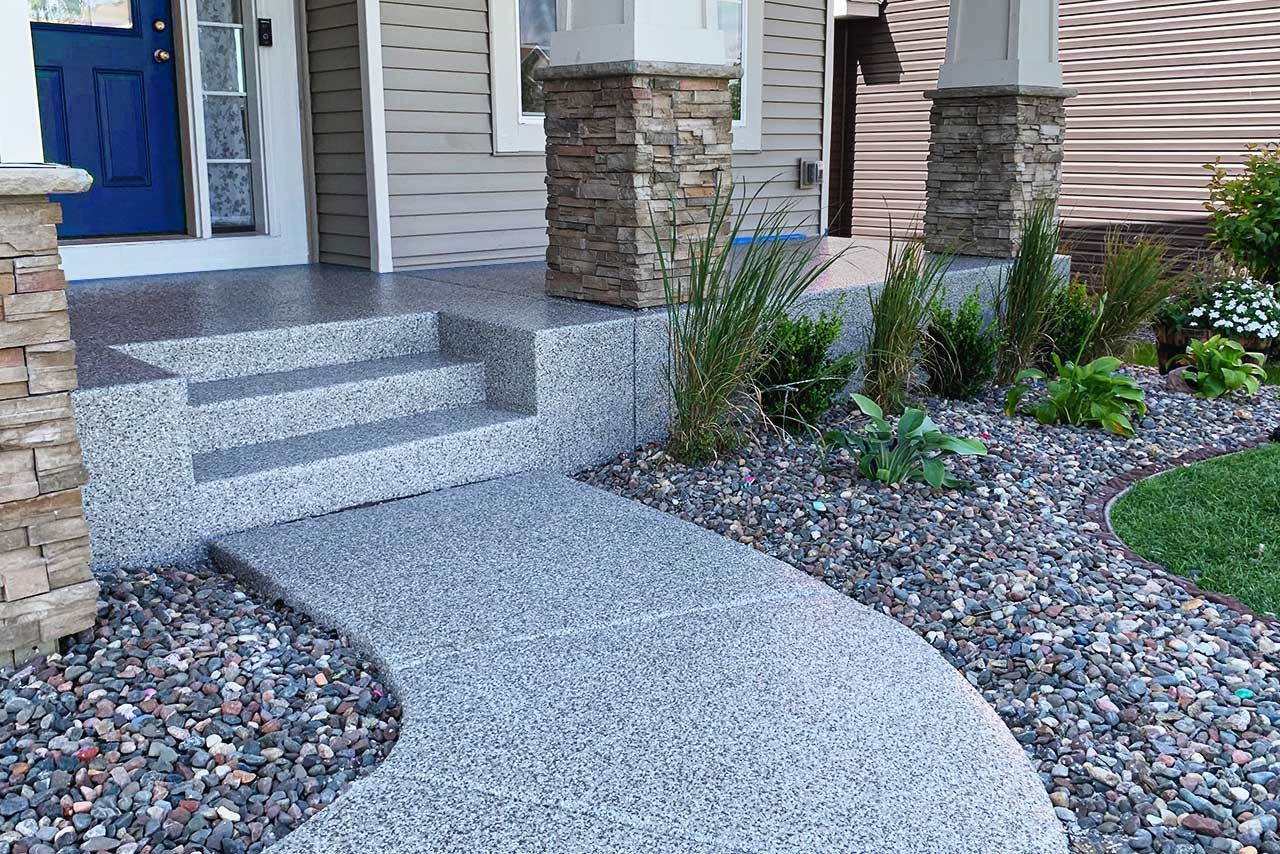 A gray speckled concrete path and steps lead to a home's blue door. Landscaping surrounds the pathway.