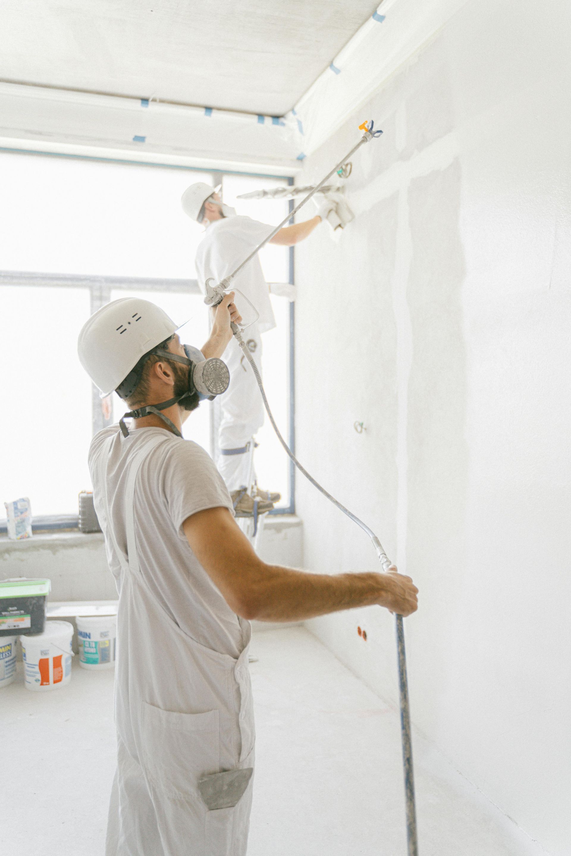 Two workers in white overalls spraying paint in a room. One on a ladder, the other holding a hose.
