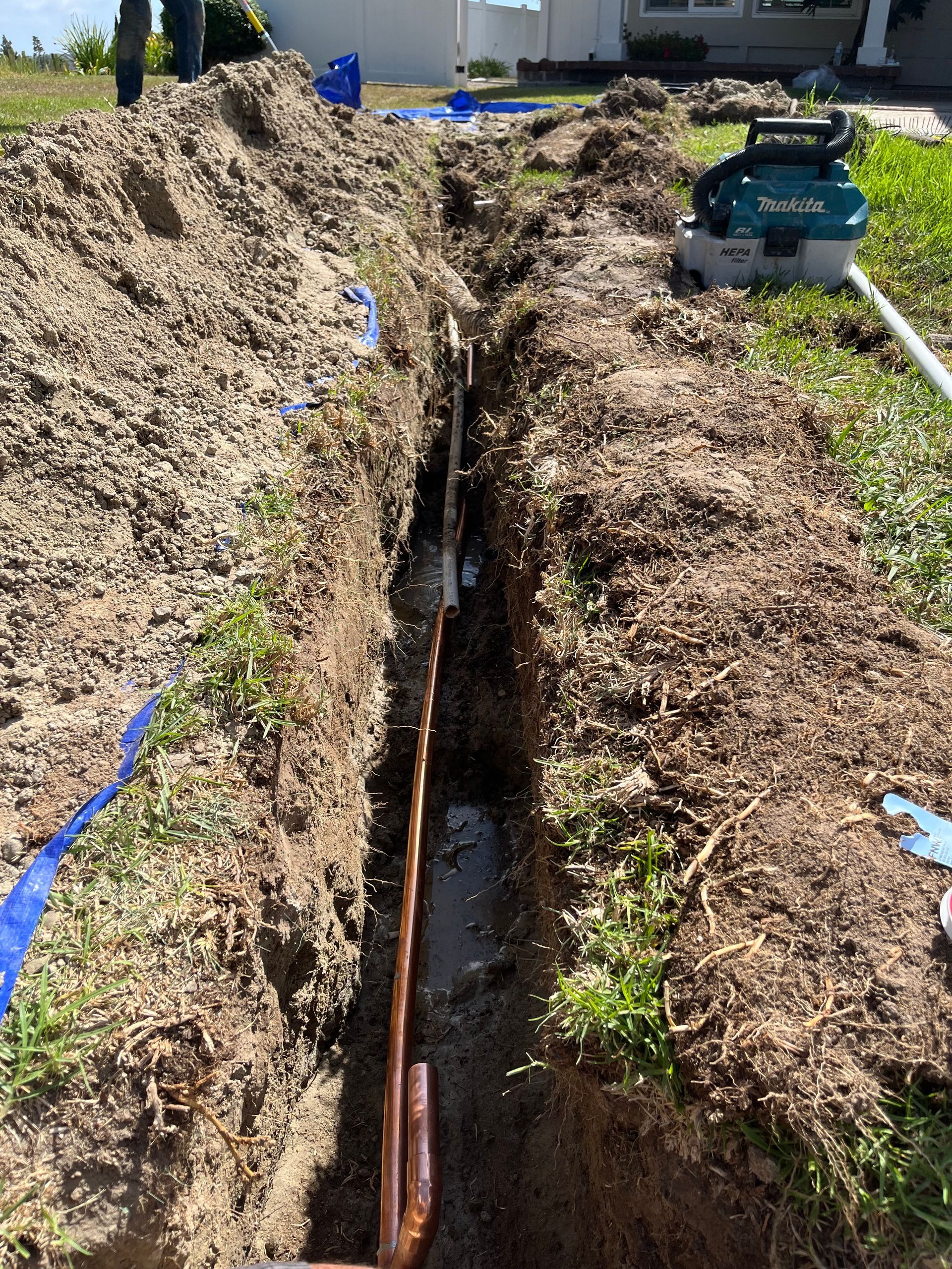 Trench dug in yard with copper piping and power tools. Blue tape marks pipe runs.