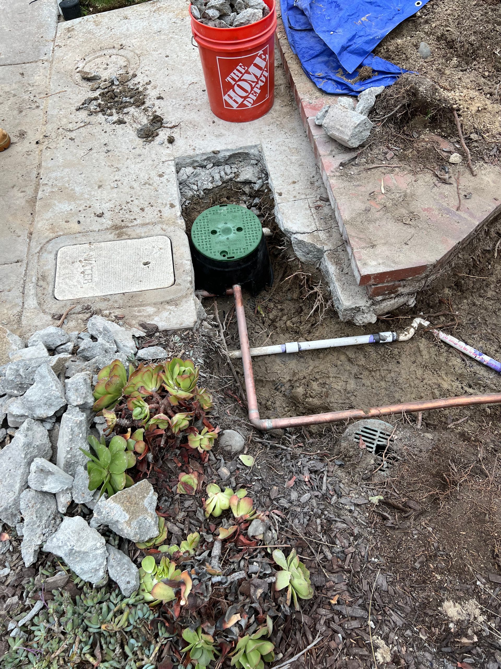 Irrigation system installation: Green valve box, copper pipes in a trench, concrete, rocks, succulents.