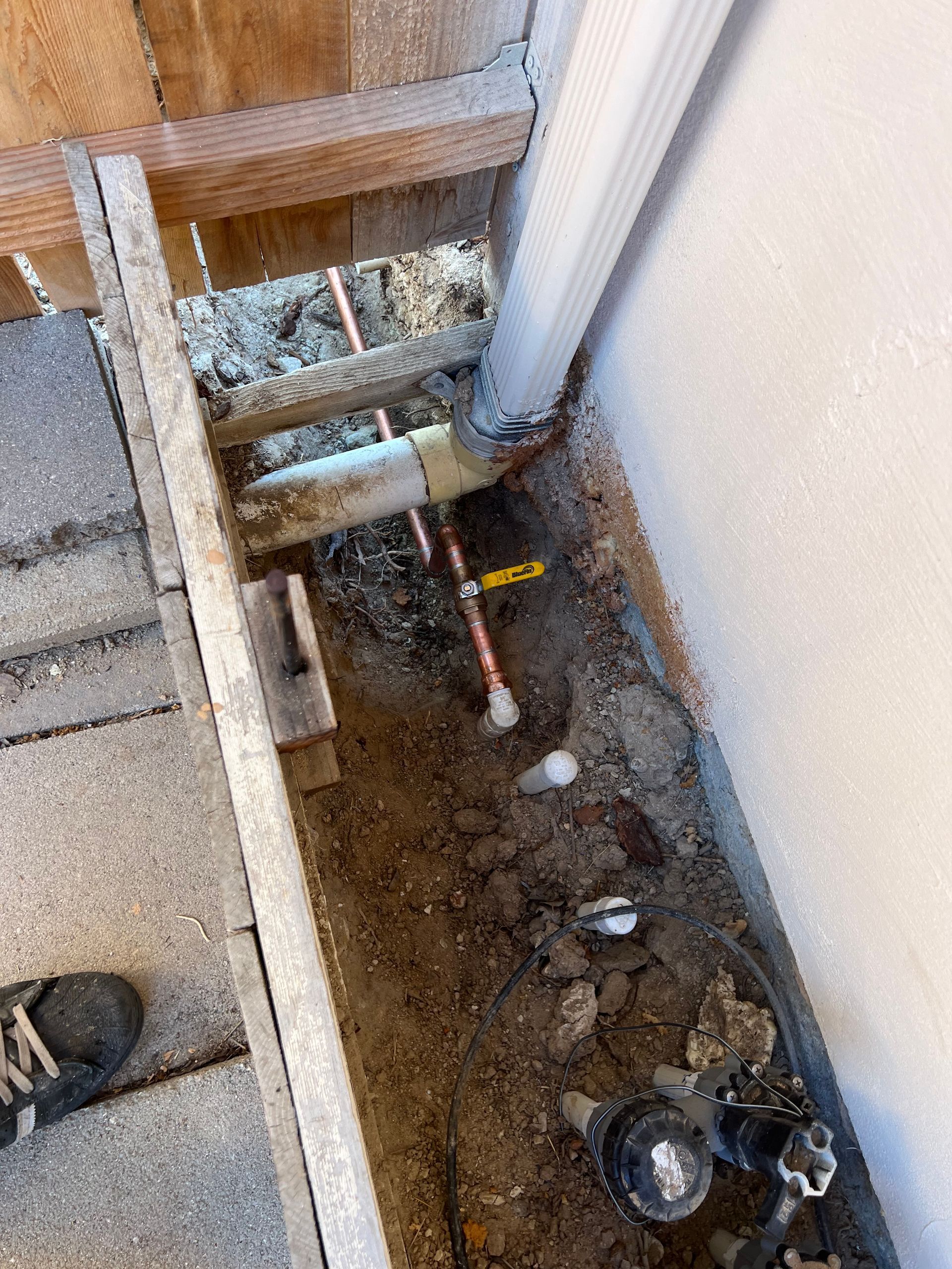 Trench with exposed copper pipes and plumbing fixtures near a building foundation.
