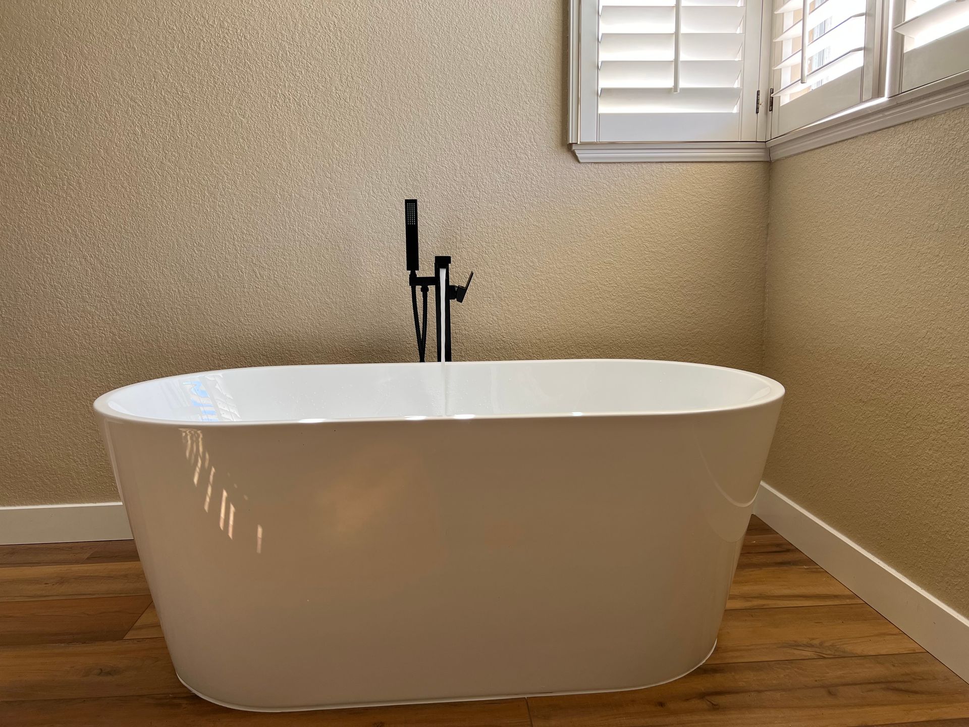 Freestanding white bathtub with black faucet, wood floor, and gold textured walls near a window with shutters.