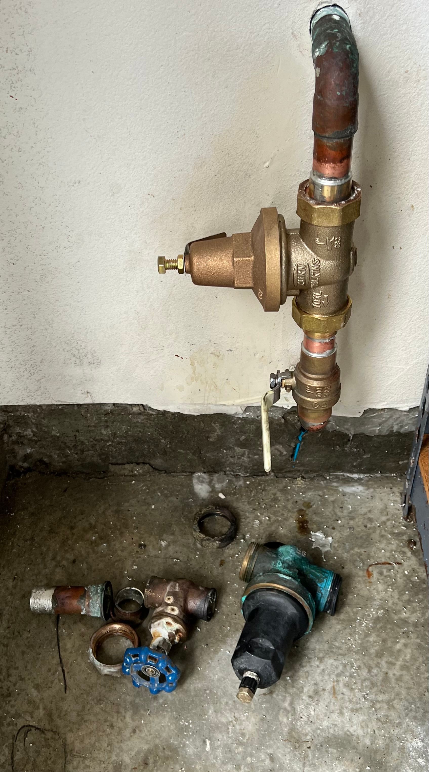 Plumbing components on a concrete floor. A pressure regulator is connected to copper pipes, and two other valves are nearby.