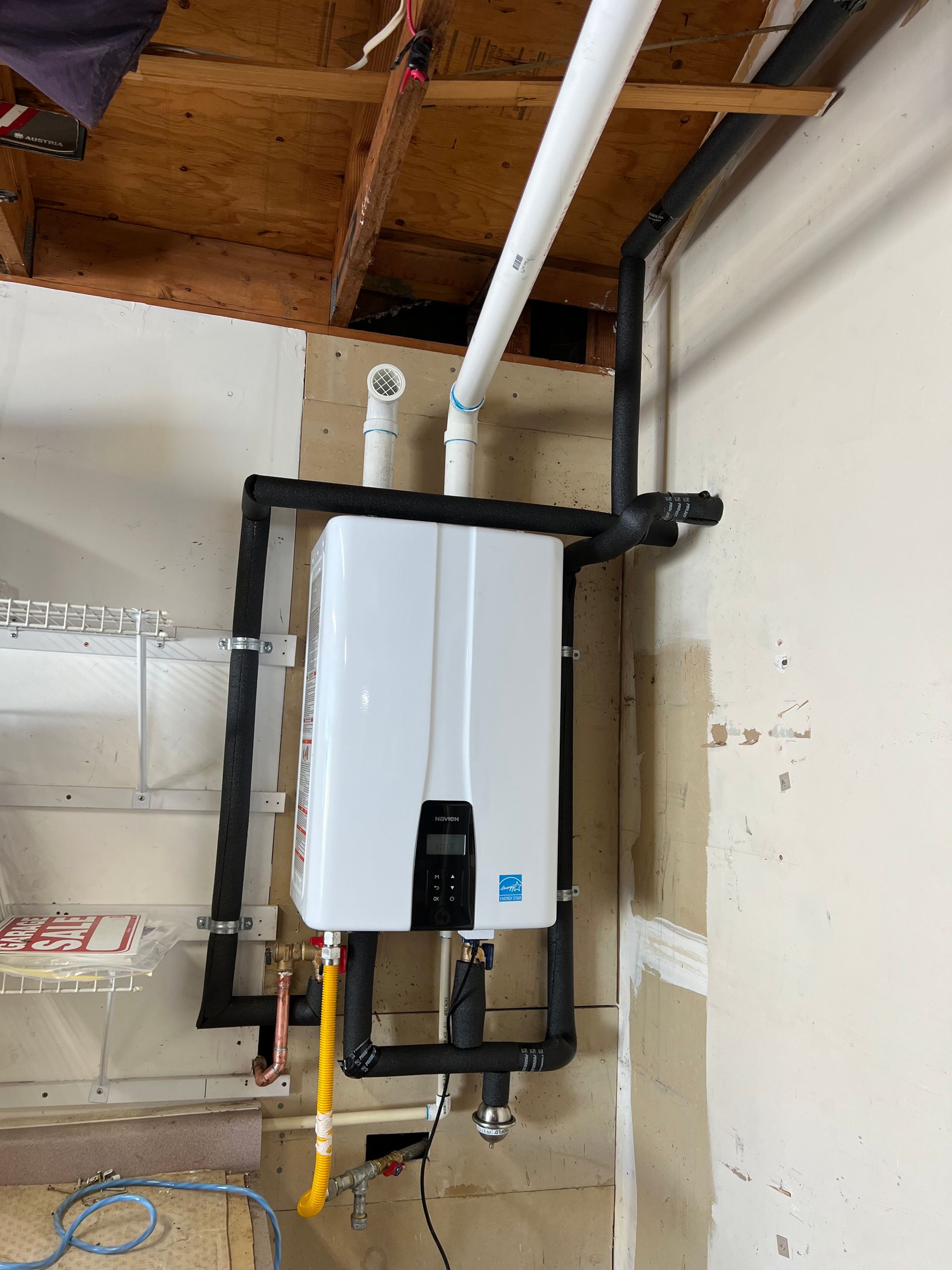A white, wall-mounted tankless water heater with black pipes and a yellow gas line in a utility room.