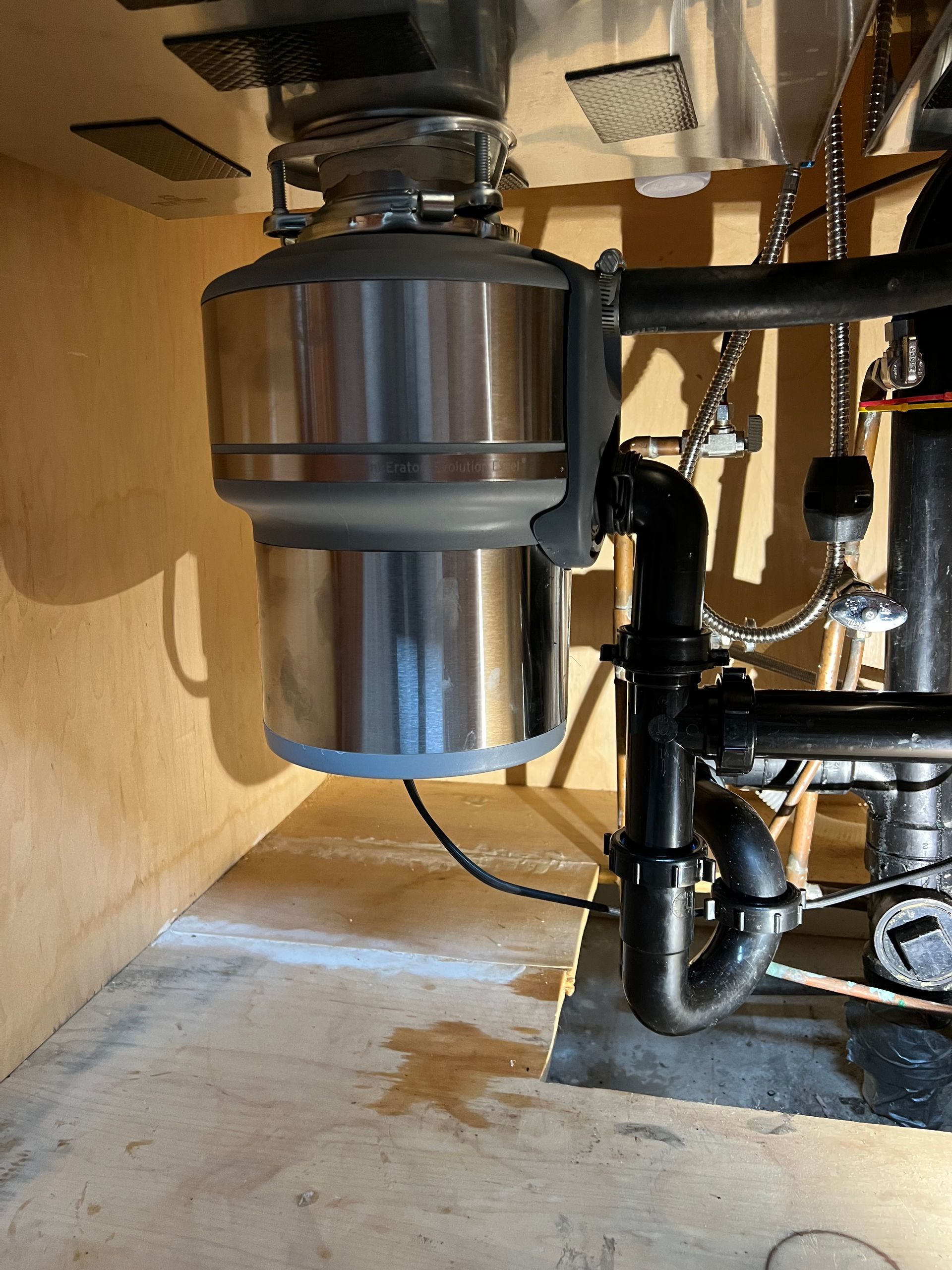 Garbage disposal unit under a kitchen sink, with black plumbing and wood cabinetry.