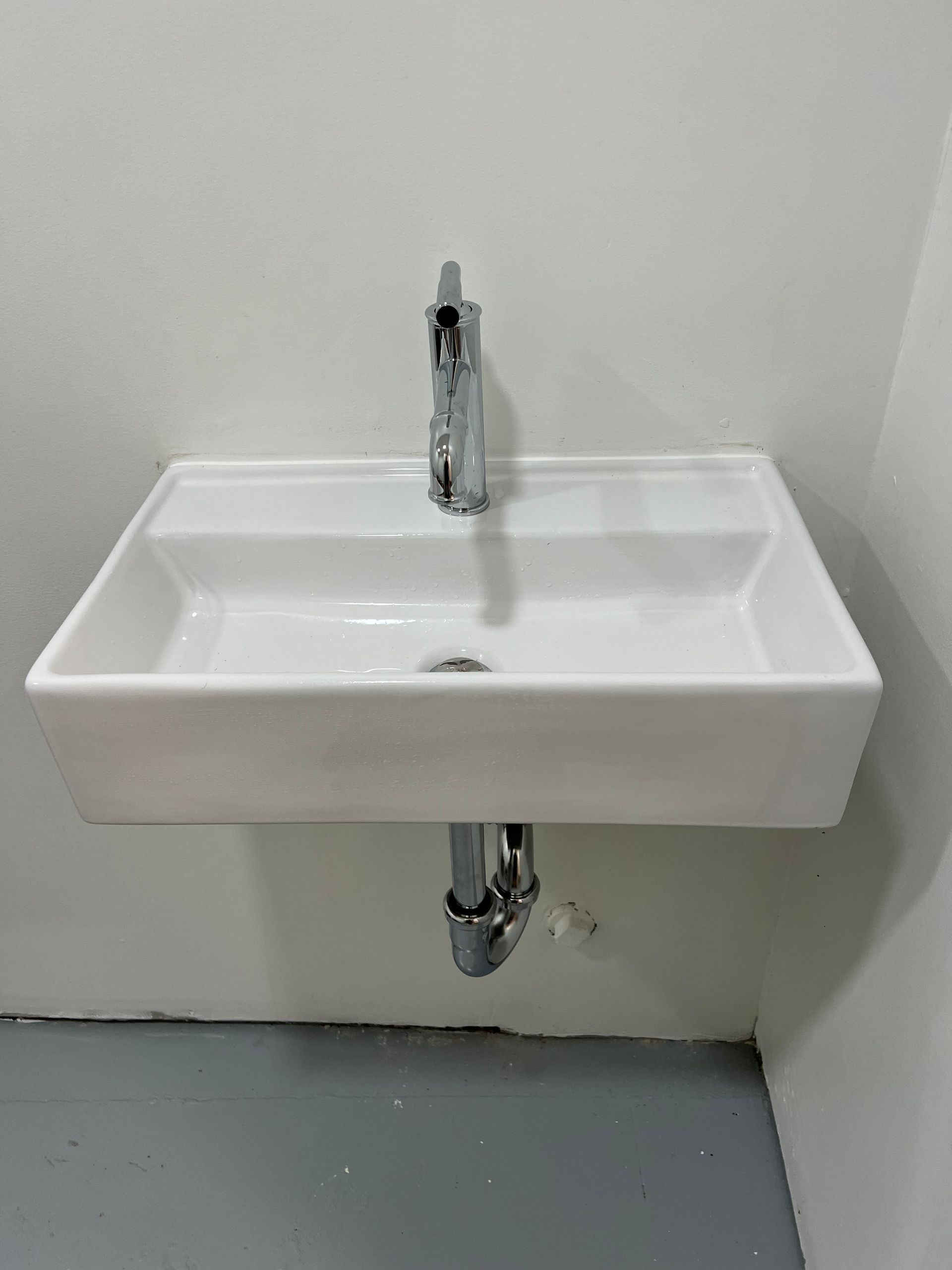 White rectangular wall-mounted sink with chrome faucet and plumbing. Grey floor, white walls.