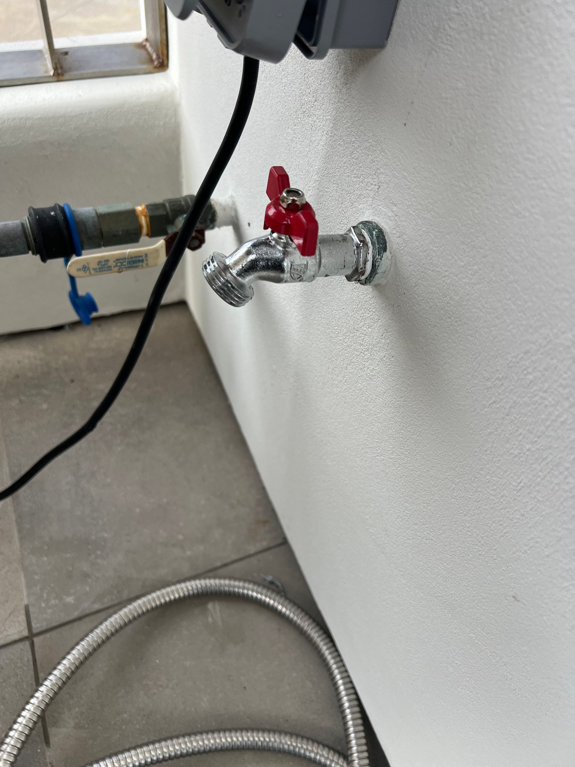 Silver faucet with red handle on white wall, connected to a hose.