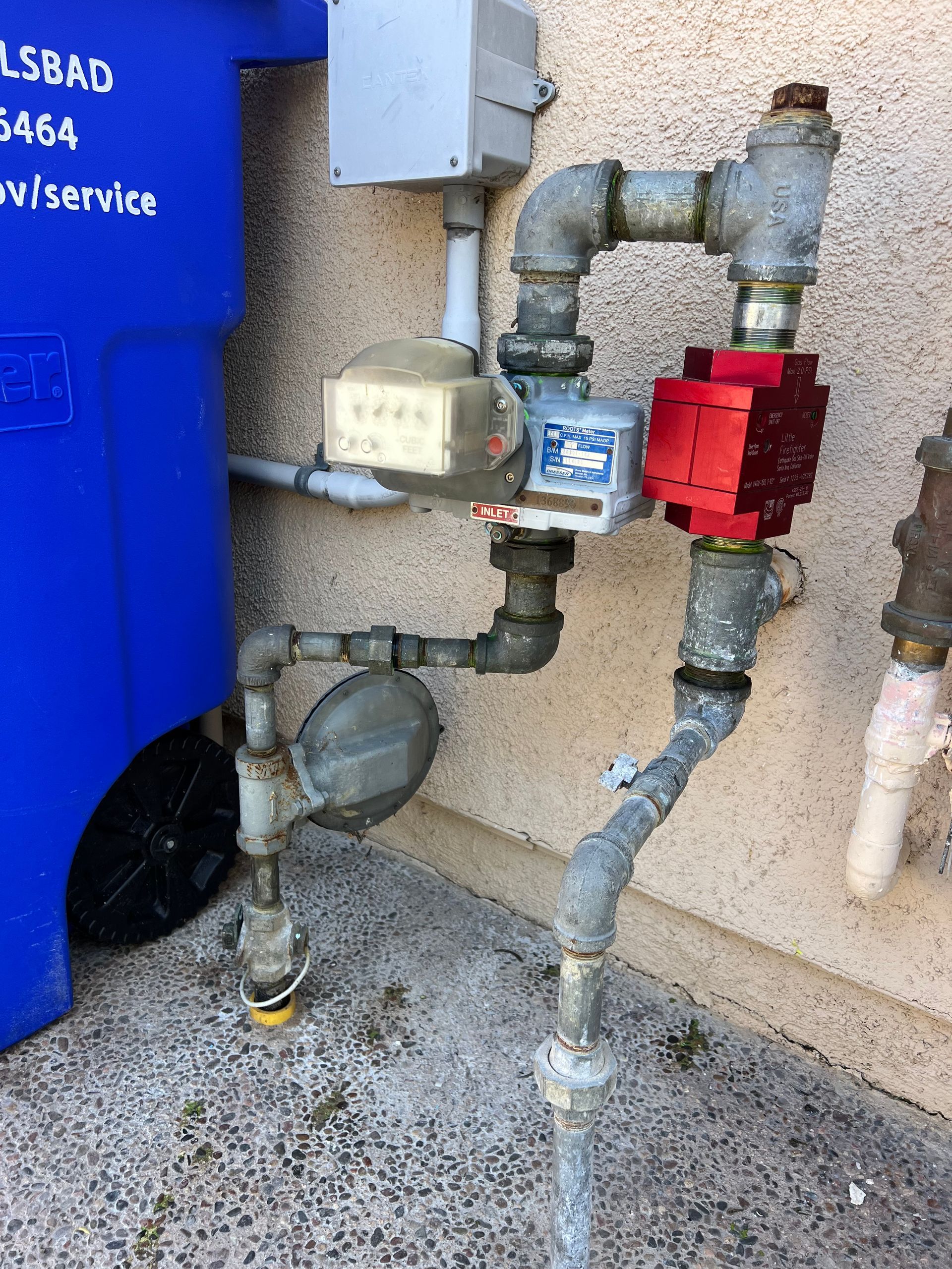 Gas meter assembly attached to a wall next to a blue trash bin. Gray pipes, a red cover, and a white meter.