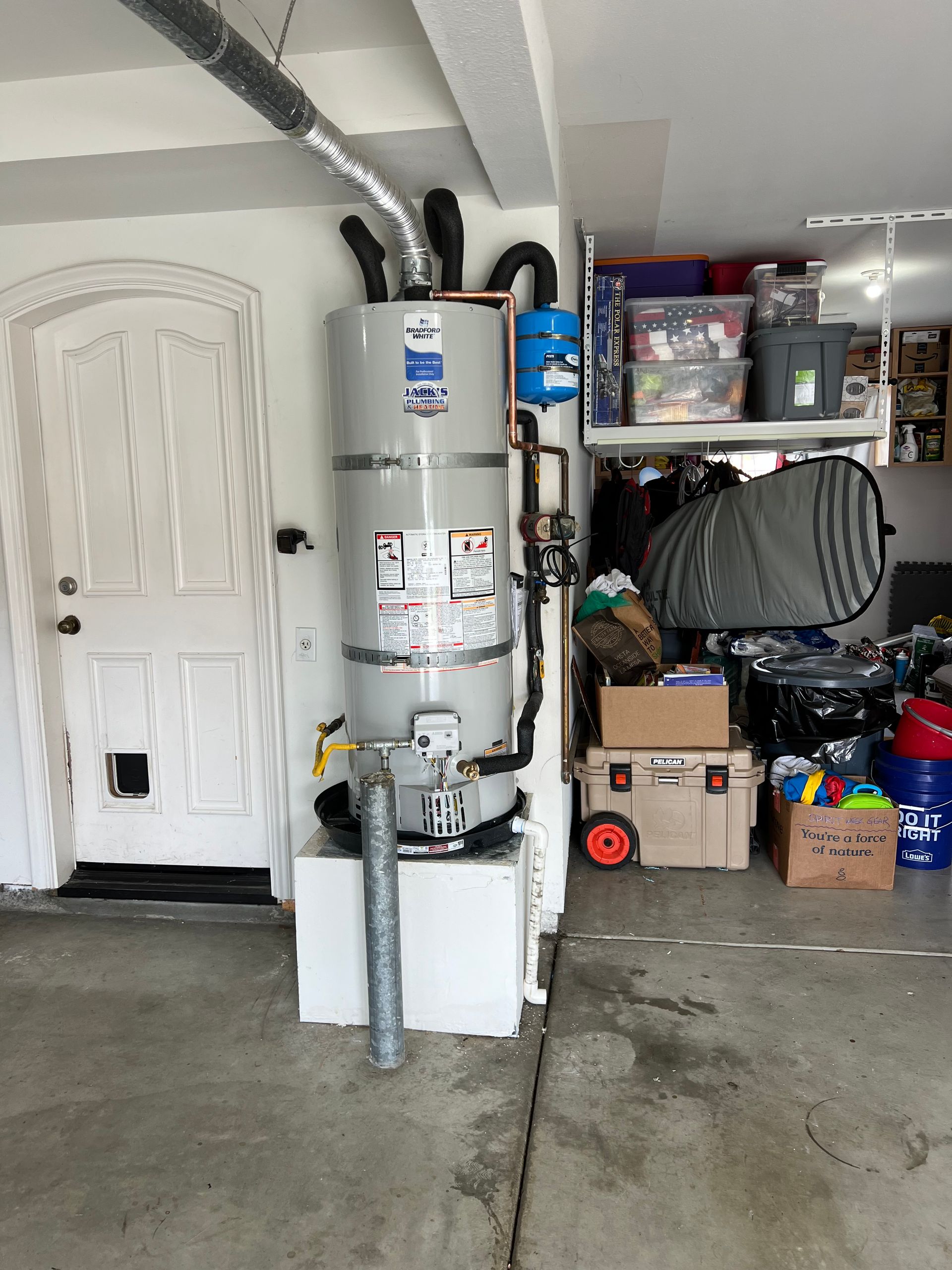 Water heater in a garage next to a white door, shelves with storage, and assorted items.