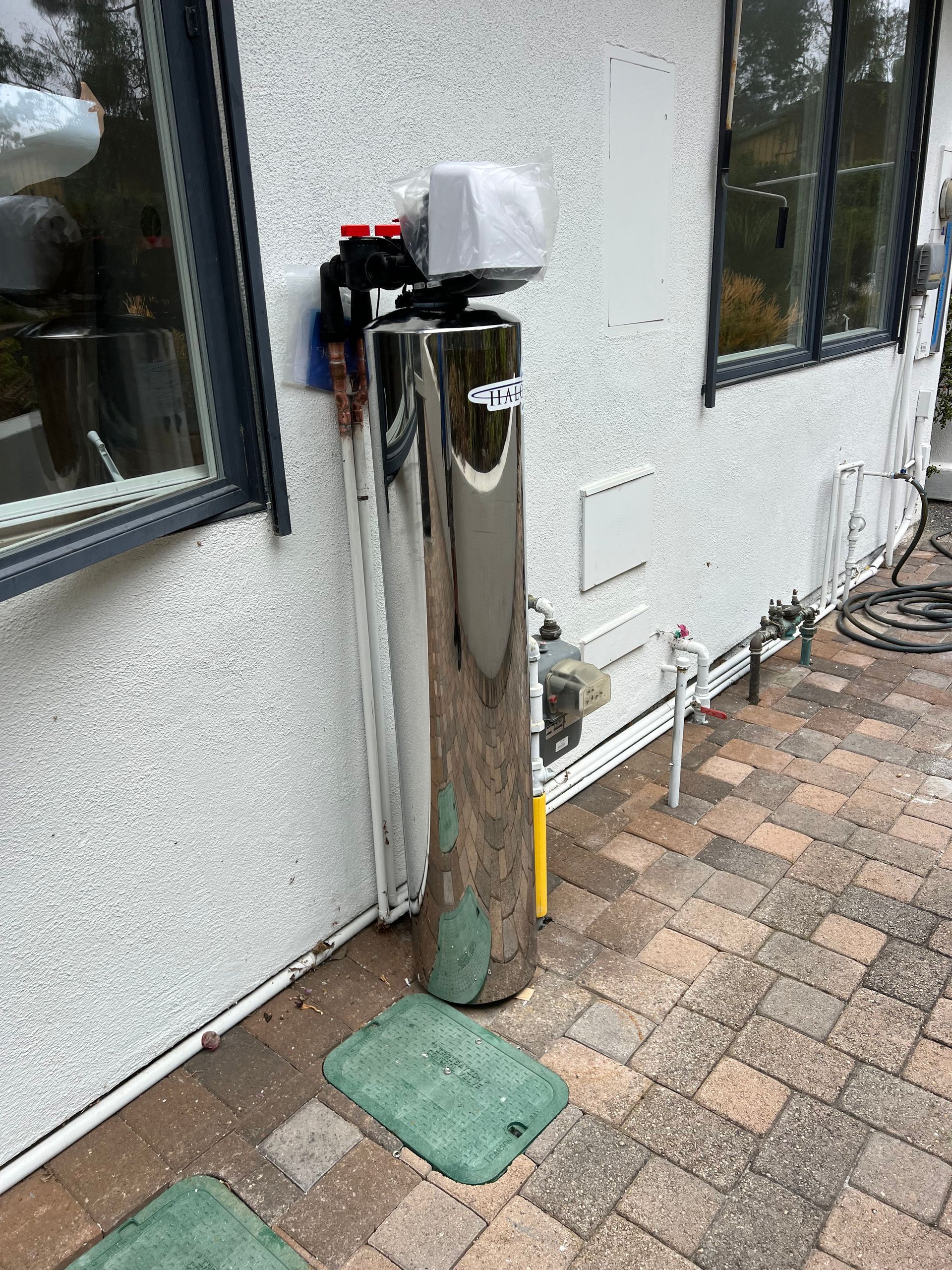 Water filtration system installed on a textured, white exterior wall, near windows, on a brick patio.