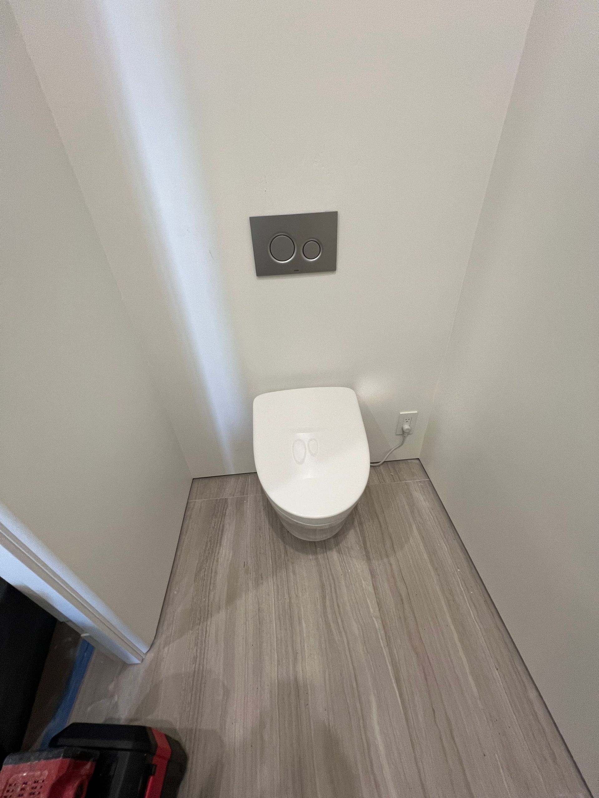 Toilet in a small bathroom with gray wood-look flooring and white walls. Flush plate is above the toilet.