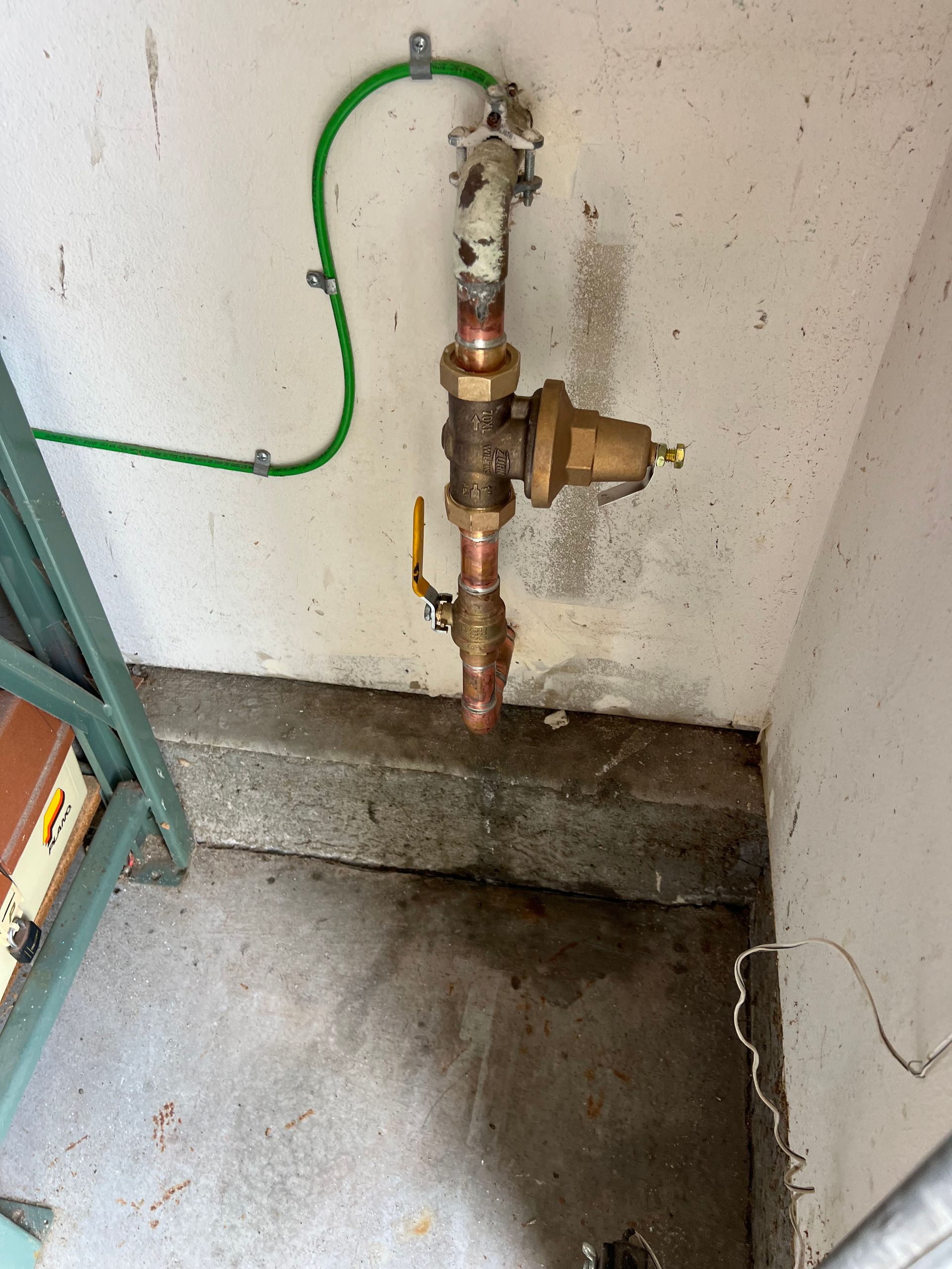 Water pressure regulator mounted on a concrete wall. Copper and brass pipes, green wire, and a yellow handle.