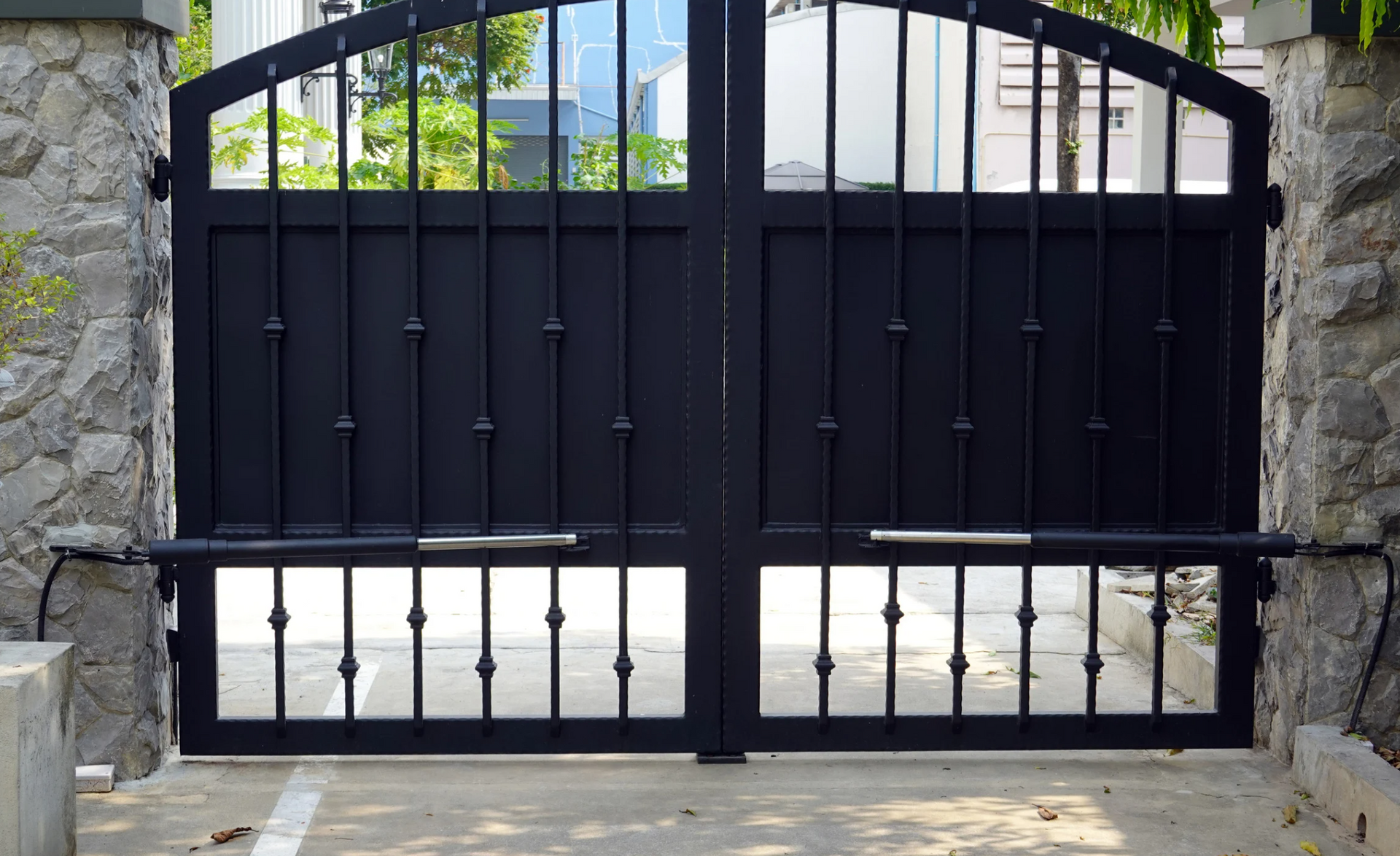 Black metal gate, closed, between stone pillars, with an arched top and bars.