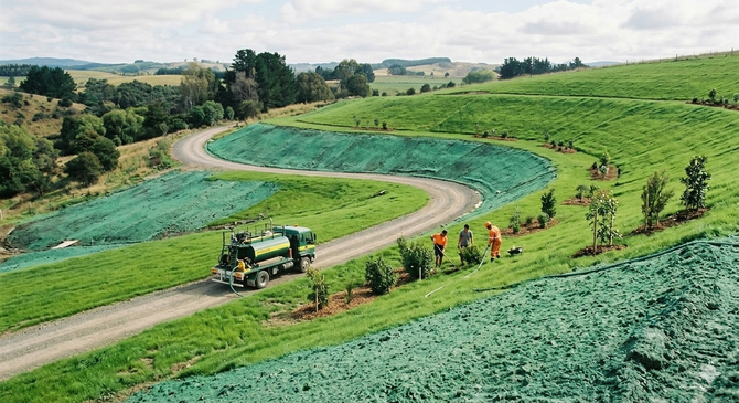 Workers in safety vests use hydroseeding trucks to spray a green mulch and seed mixture onto a dirt hillside.