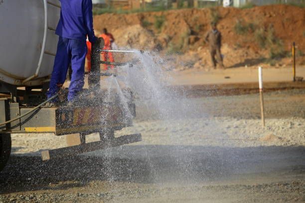 A worker sprays water from a tank truck onto a dusty construction site to dampen the ground.