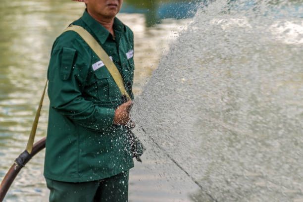 A person in a green uniform sprays a powerful stream of water from a hose near a body of water.