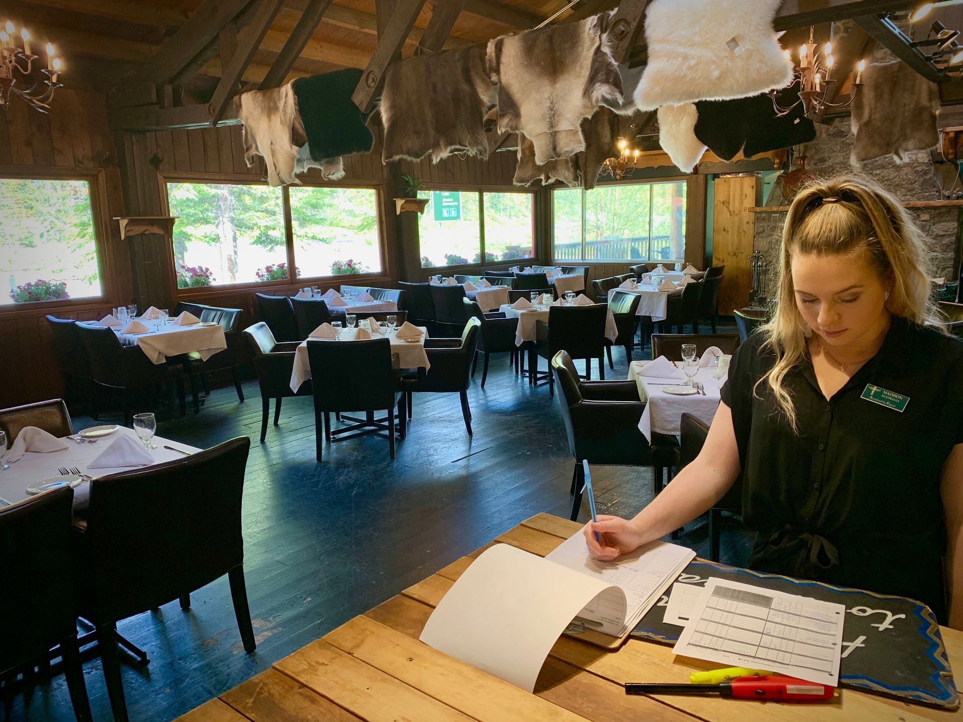 A server in a restaurant writes in a notebook at a wooden counter. Tables are set in the background.