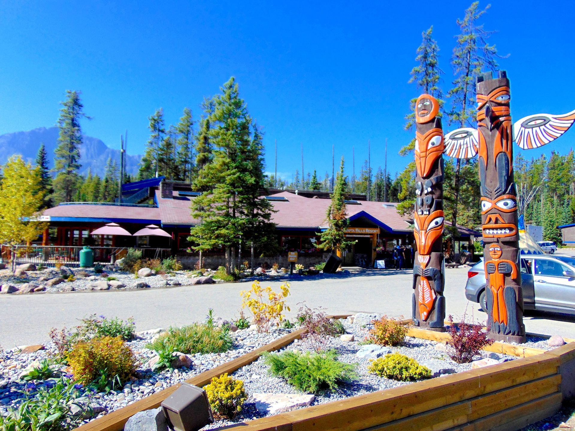 Two totem poles stand in front of a restaurant, mountains in the background. Bright blue sky.